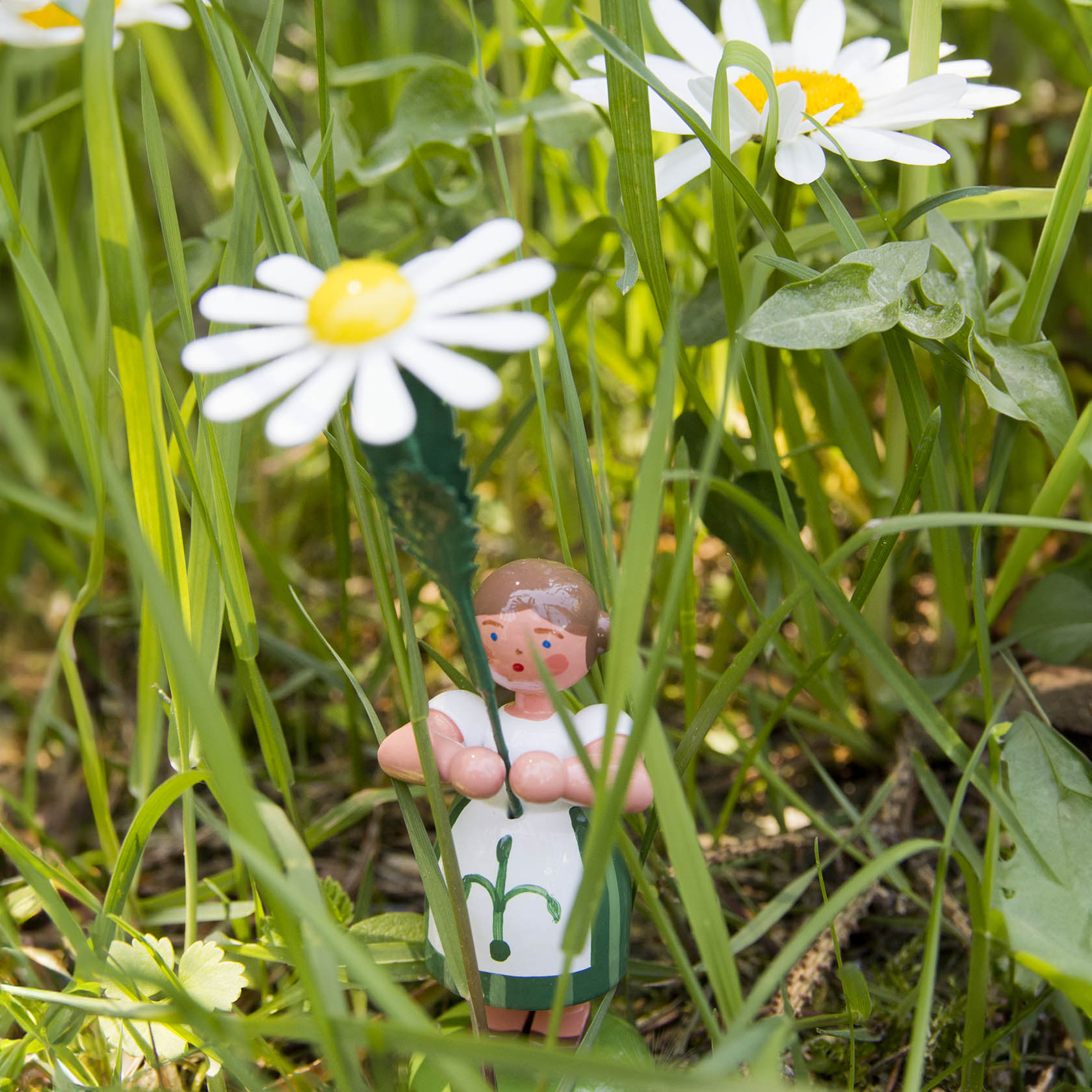 Eine kleine Holzfigur eines Mädchens mit Zöpfen und grünem Kleid hält eine große Margerite in einer Wiese.