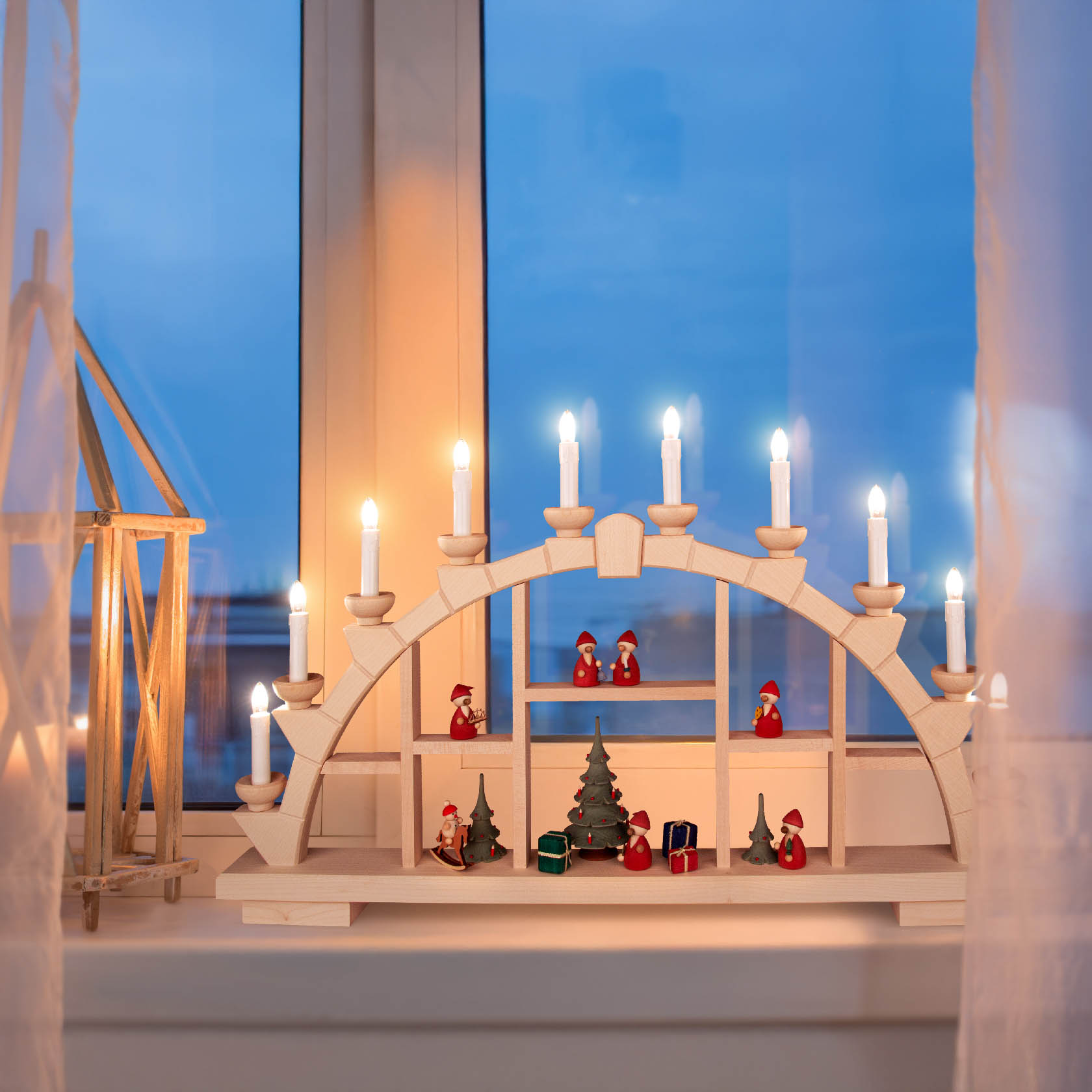 Beleuchteter Schwibbogen mit Setzkasten, aus hellem Holz, auf Fensterbank. Mehrere Kerzen auf dem Bogen, darunter Weihnachtsdekoration wie kleine Figuren und Bäume.