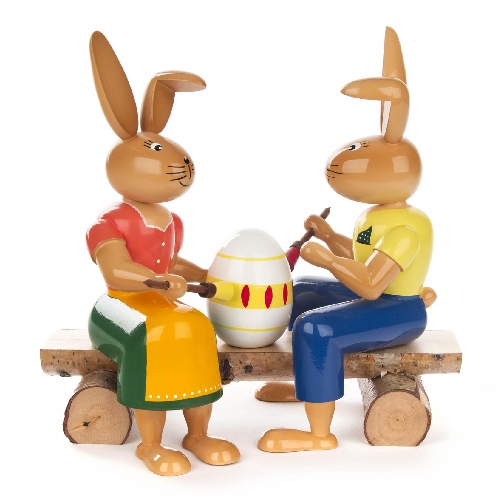 Zwei Hasen mit Malutensilien, die auf einer Holzbank sitzen und ein großes Osterei bemalen. Einer der Hasen trägt ein rotes Oberteil, der andere ein gelbes. im Dregeno Online Shop günstig kaufen