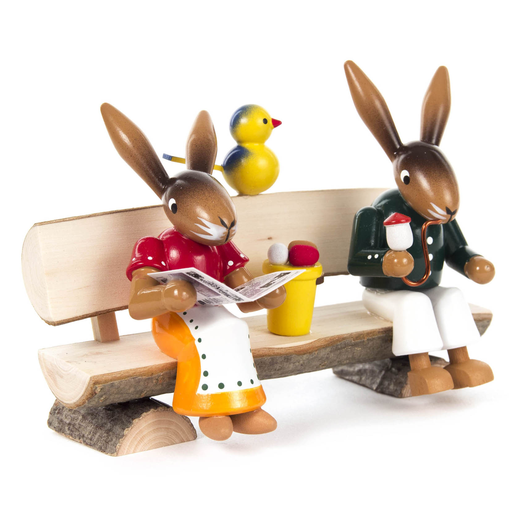 Zwei Hasenfiguren sitzen auf einer Holzbank. Einer hält eine Zeitung, der andere eine Tasse. Ein gelber Vogel sitzt auf der Rückenlehne.