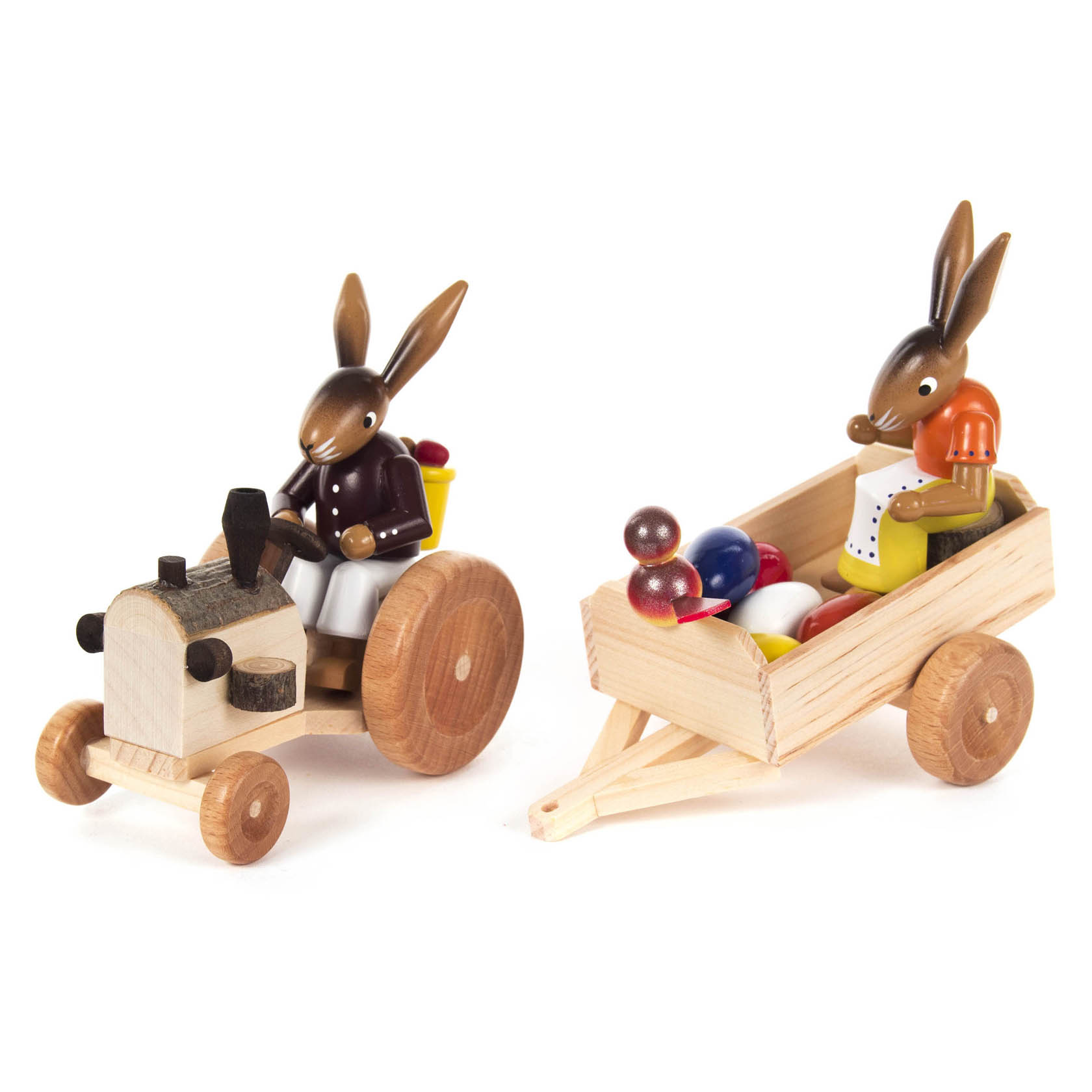 Zwei hölzerne Hasenfiguren, eine auf einem Traktor mit gelober Schaufel hinten und die andere auf einem Anhänger sitzend. Der Anhänger ist mit bunten Eiern gefüllt.