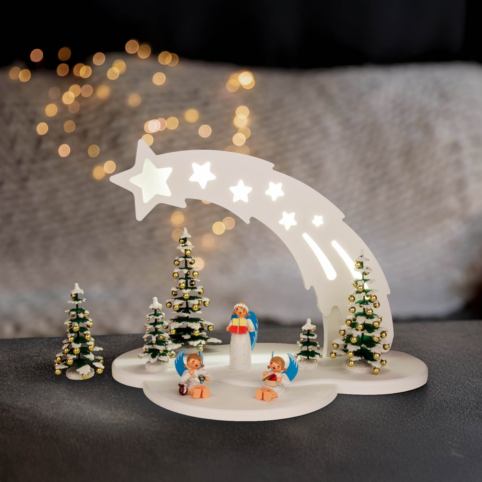 Ein weißer Sternbogen mit LED-Beleuchtung, darunter Engel auf Vorlegern. Drei geschmückte Miniatur-Weihnachtsbäume umgeben die Szene.