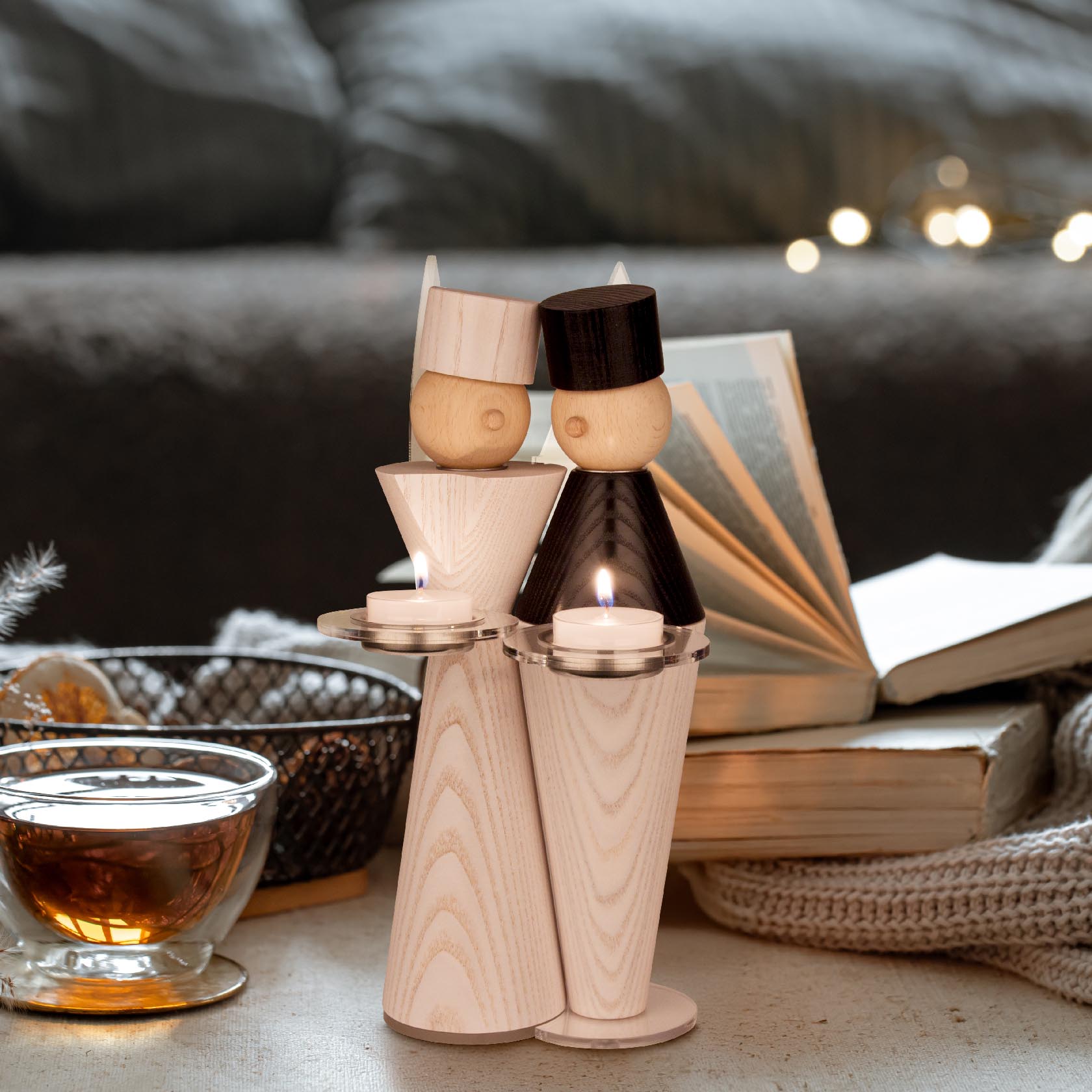 Two wooden miner figures holding candles, one with a black hat and coat, the other with a natural wood finish, on a cozy setup with tea and an open book.