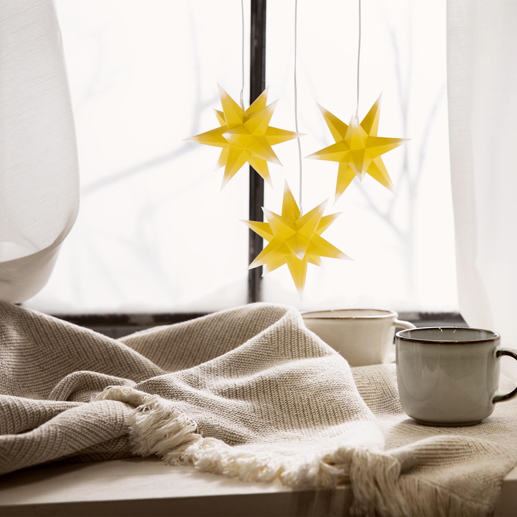 Drei leuchtende, gelbe Adventssterne mit weißen Spitzen hängen vor einem Fenster. Darunter liegt eine beige Decke neben zwei grauen Tassen.