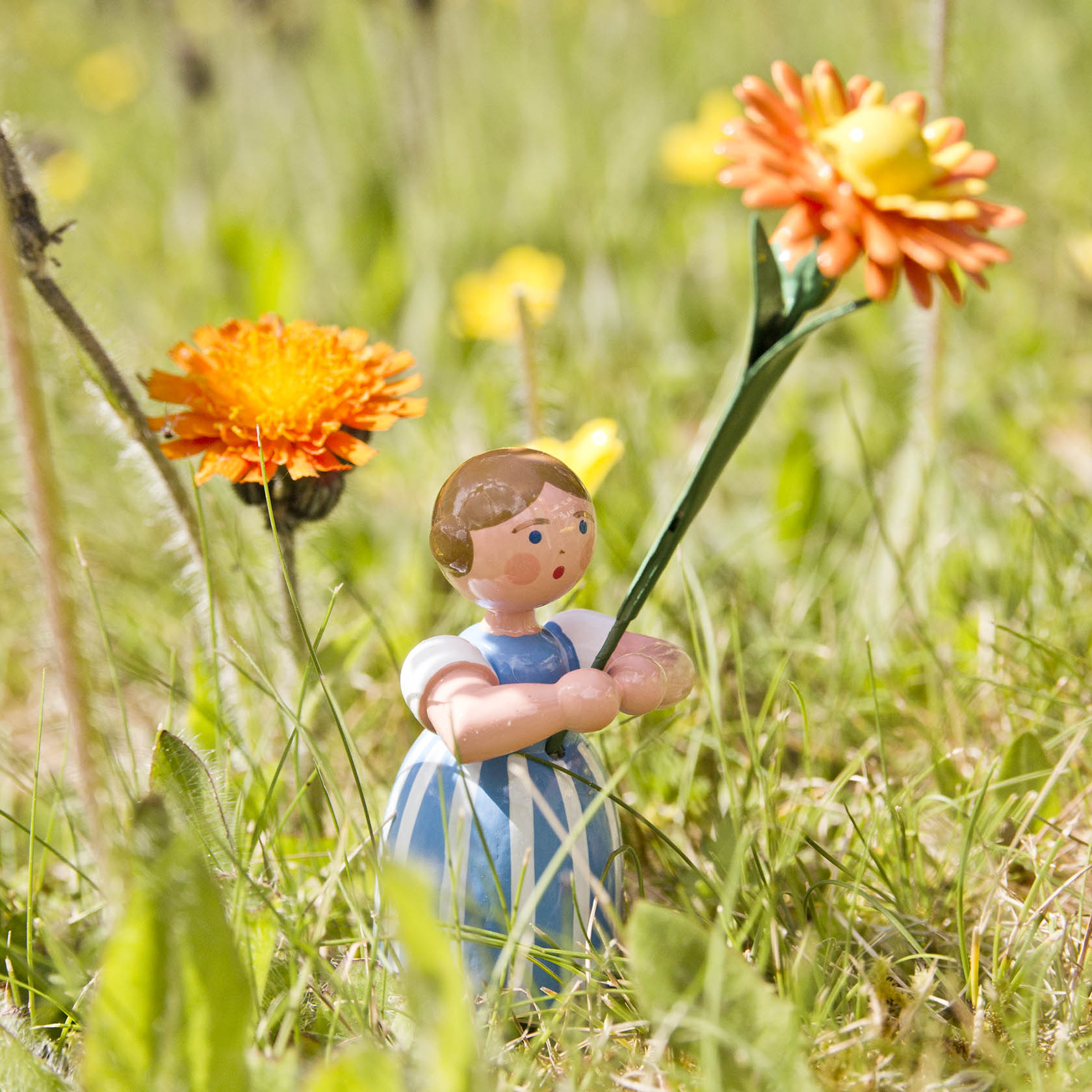 Kleine Keramikfigur eines Mädchens in einem blauen Kleid mit weißen Ärmeln, das eine große Ringelblume hält, umgeben von weiteren Ringelblumen im Gras.