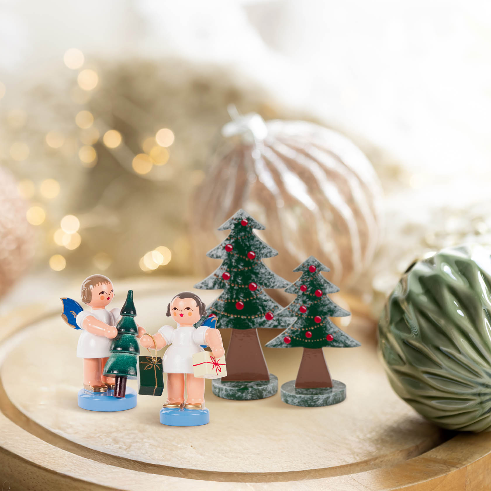 Zwei Engel-Figuren mit Flügeln, einer hält einen kleinen Weihnachtsbaum, der andere ein Geschenk. Drei kleine, flache Weihnachtsbaumfiguren stehen daneben.