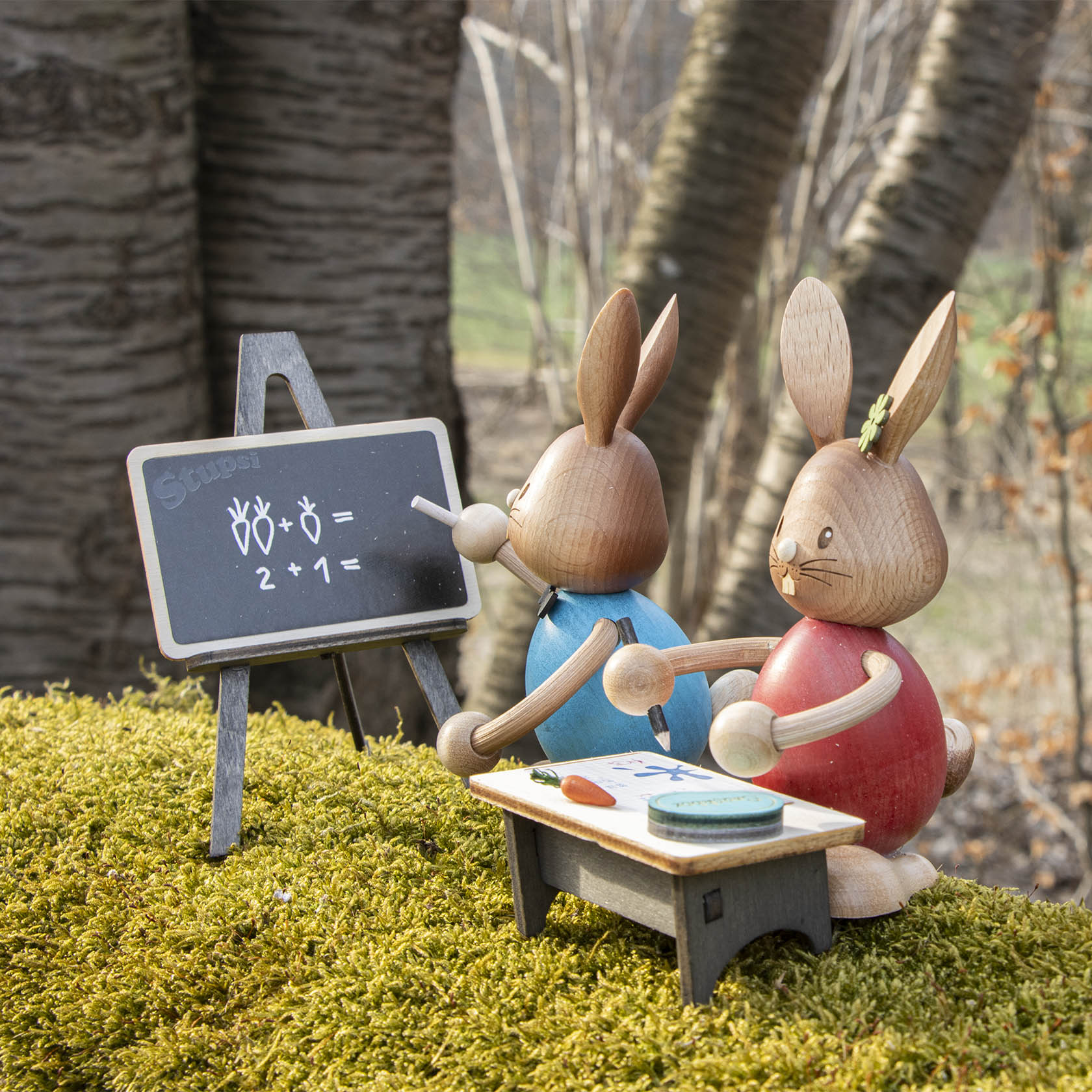 Zwei Holzhasen, einer mit Kreide vor einer kleinen Tafel stehend, auf der einfache Mathematikaufgaben geschrieben sind. Der andere Hase sitzt an einem Schreibtisch.