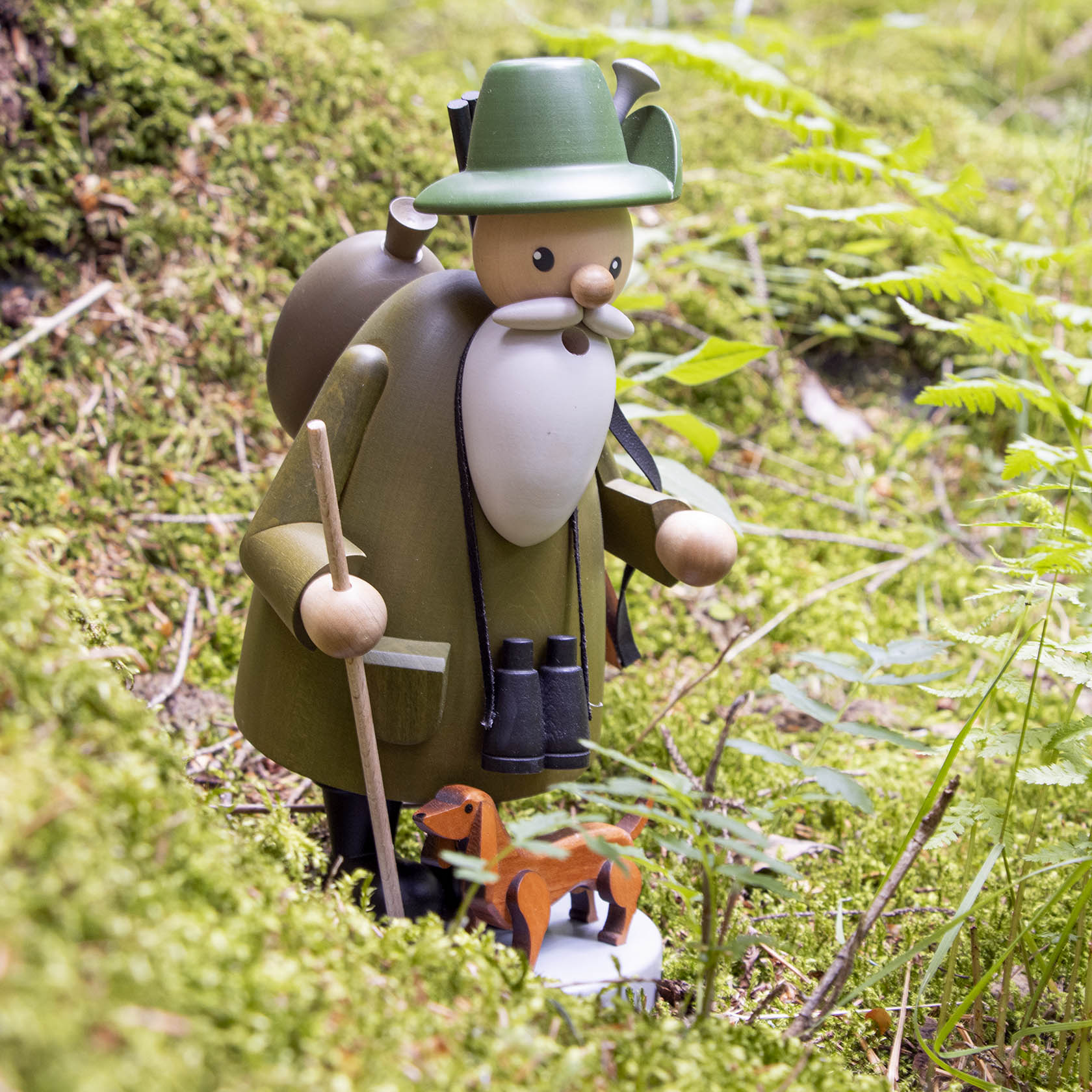 Holzfigur eines Försters mit grünem Hut und Mantel, trägt Fernglas und Spazierstock; ein kleiner Dackel aus Holz steht neben ihm.