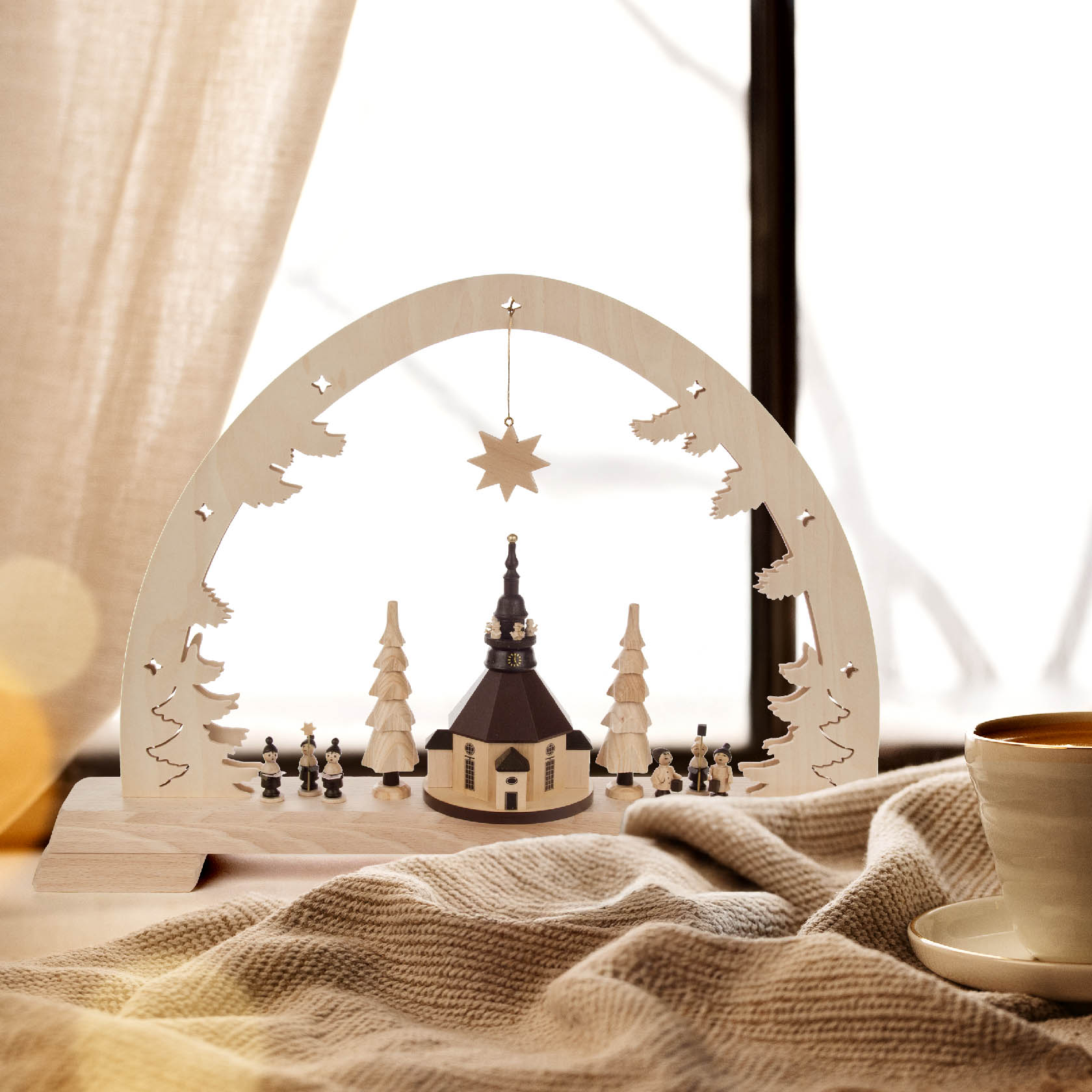 Ein Schwibbogen mit einer Miniatur der Seiffener Kirche, umgeben von LED-beleuchteten Tannenbäumen und Figuren, darunter ein Stern. Im Vordergrund eine Tasse.