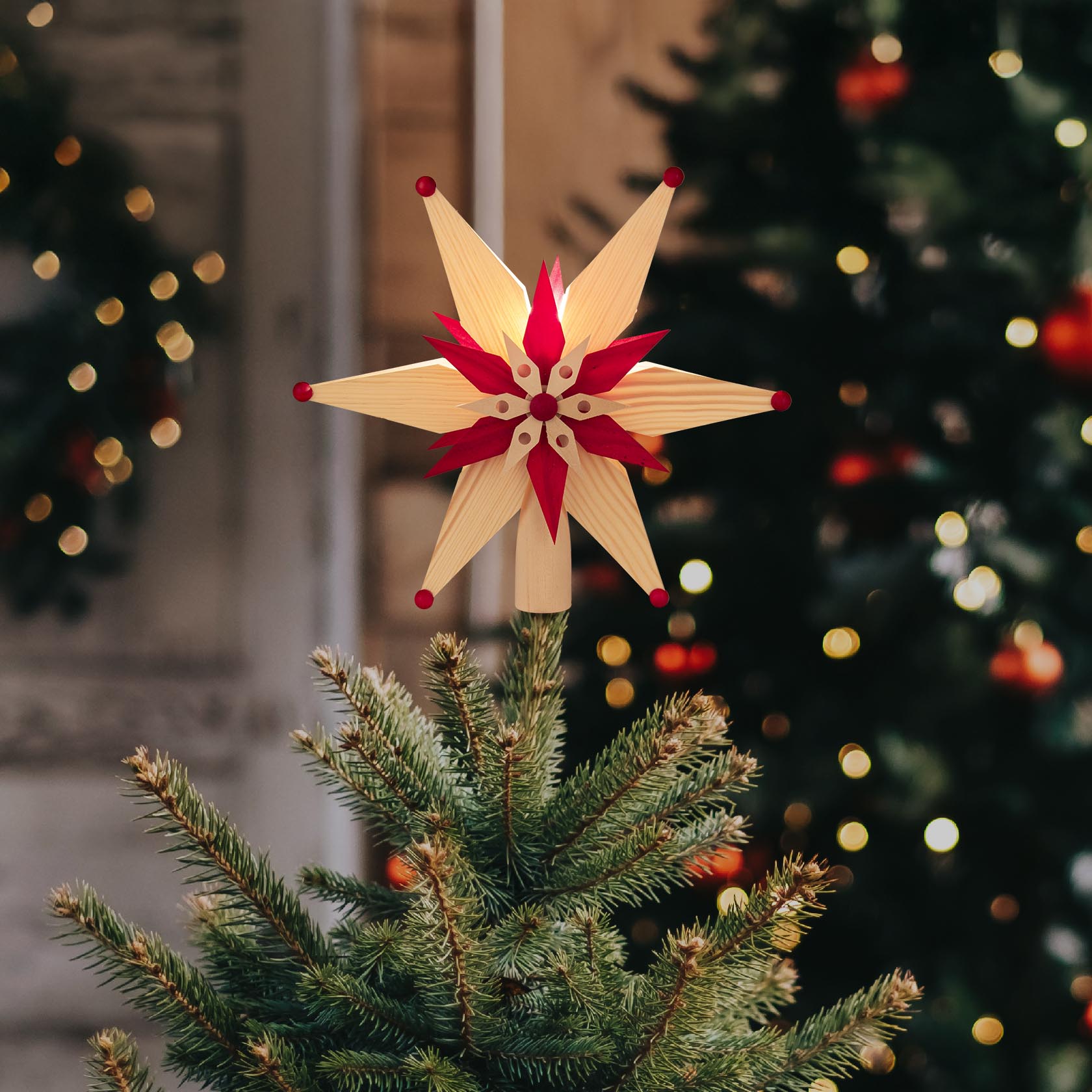 Eine Christbaumspitze in Form eines Spansterns, in Rot und Natur gehalten, mit elektrischer Beleuchtung, auf einem Tannenbaum angebracht.