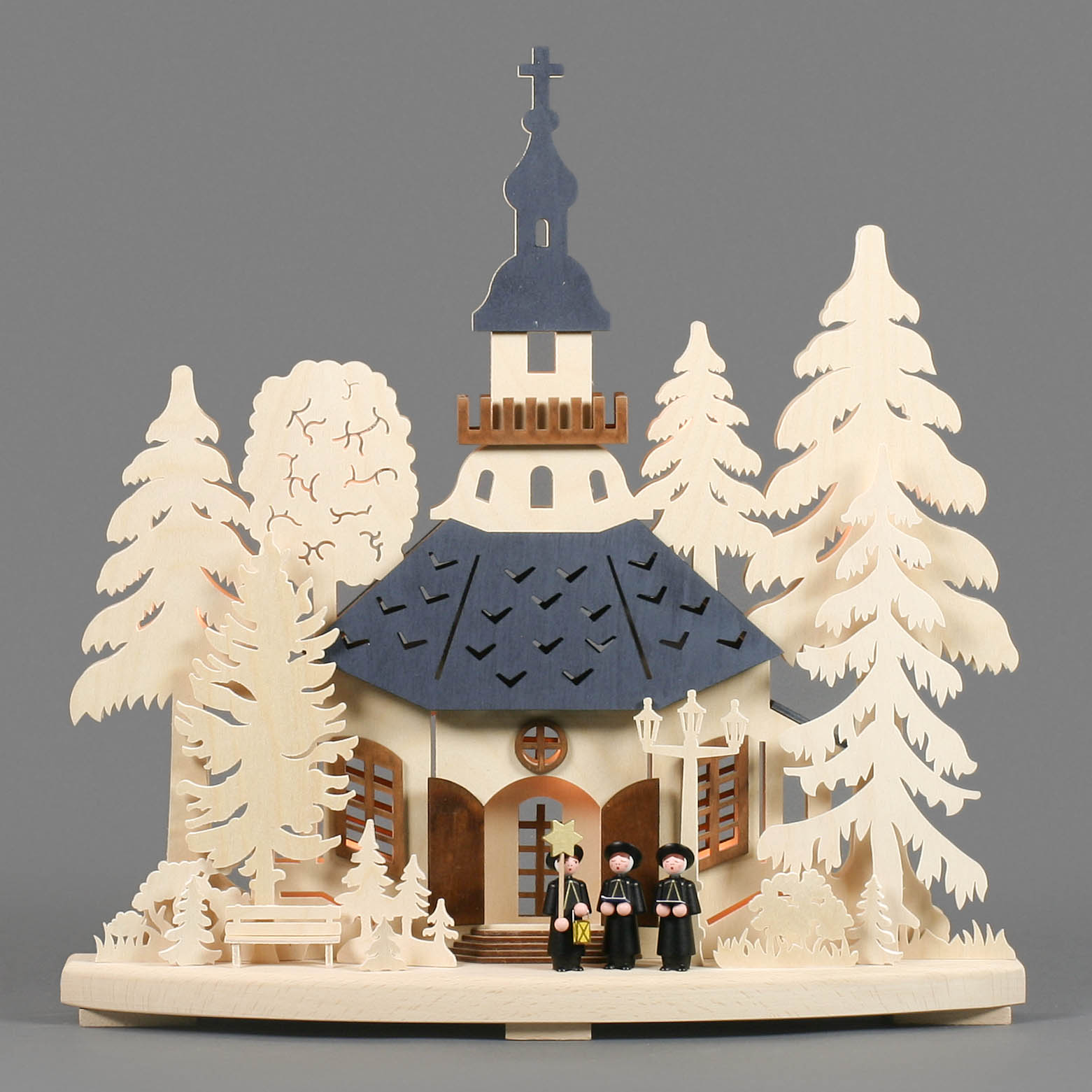 Schwibbogen aus Holz mit Seiffener Kirche und drei Figuren davor, elektrisch beleuchtet. Kirche hat einen dunkelblauen Turm, umgeben von großen Bäumen.