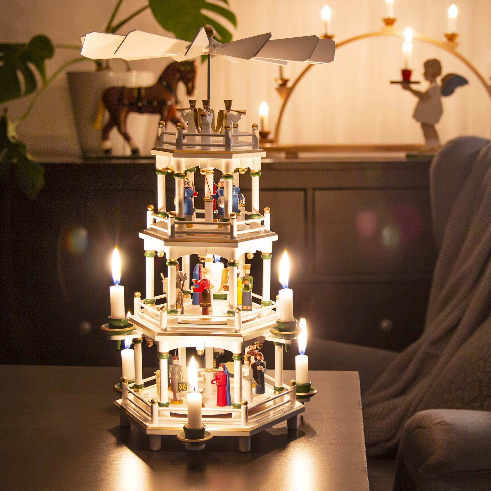 Dreistöckige weiße Weihnachtspyramide mit Szenen der Christi Geburt, angetrieben von Kerzen; Figuren in traditionellen Farben, darunter Maria, Josef, das Jesuskind und die Heiligen Drei Könige.
