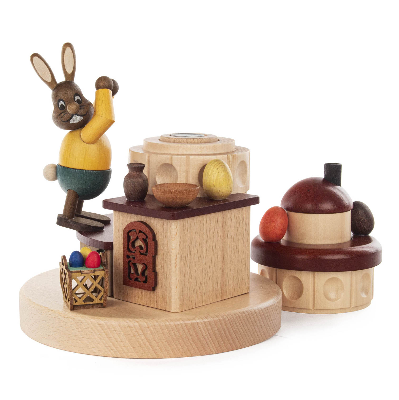 Holzfigur eines lächelnden Hasen mit erhobenen Armen neben einem Ofen, daneben ein kleiner Korb mit bunten Ostereiern.