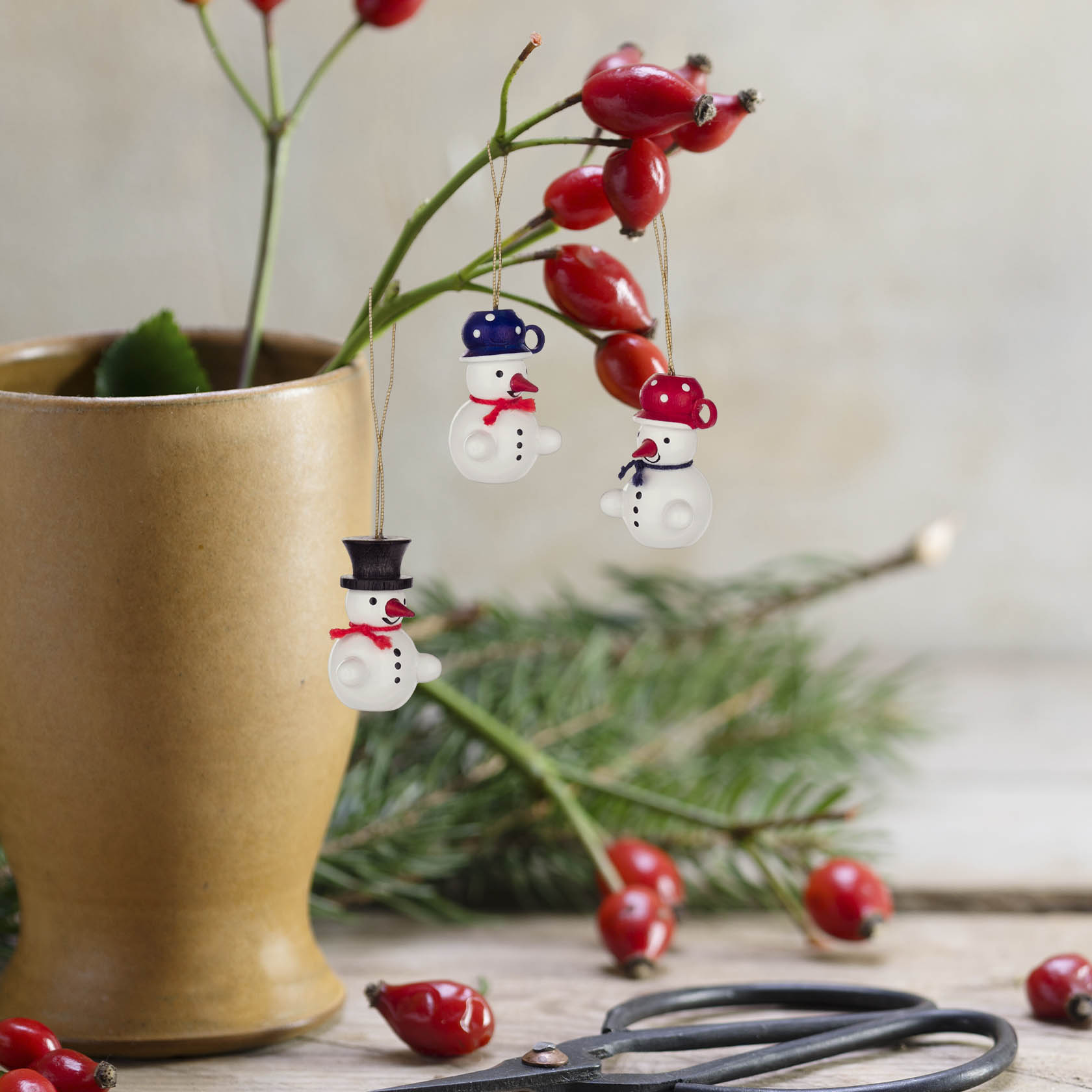 Drei hängende Schneemann-Ornamente in verschiedenen Designs an Zweigen mit roten Beeren, platziert in einer Vase vor einem Ast mit Nadeln.