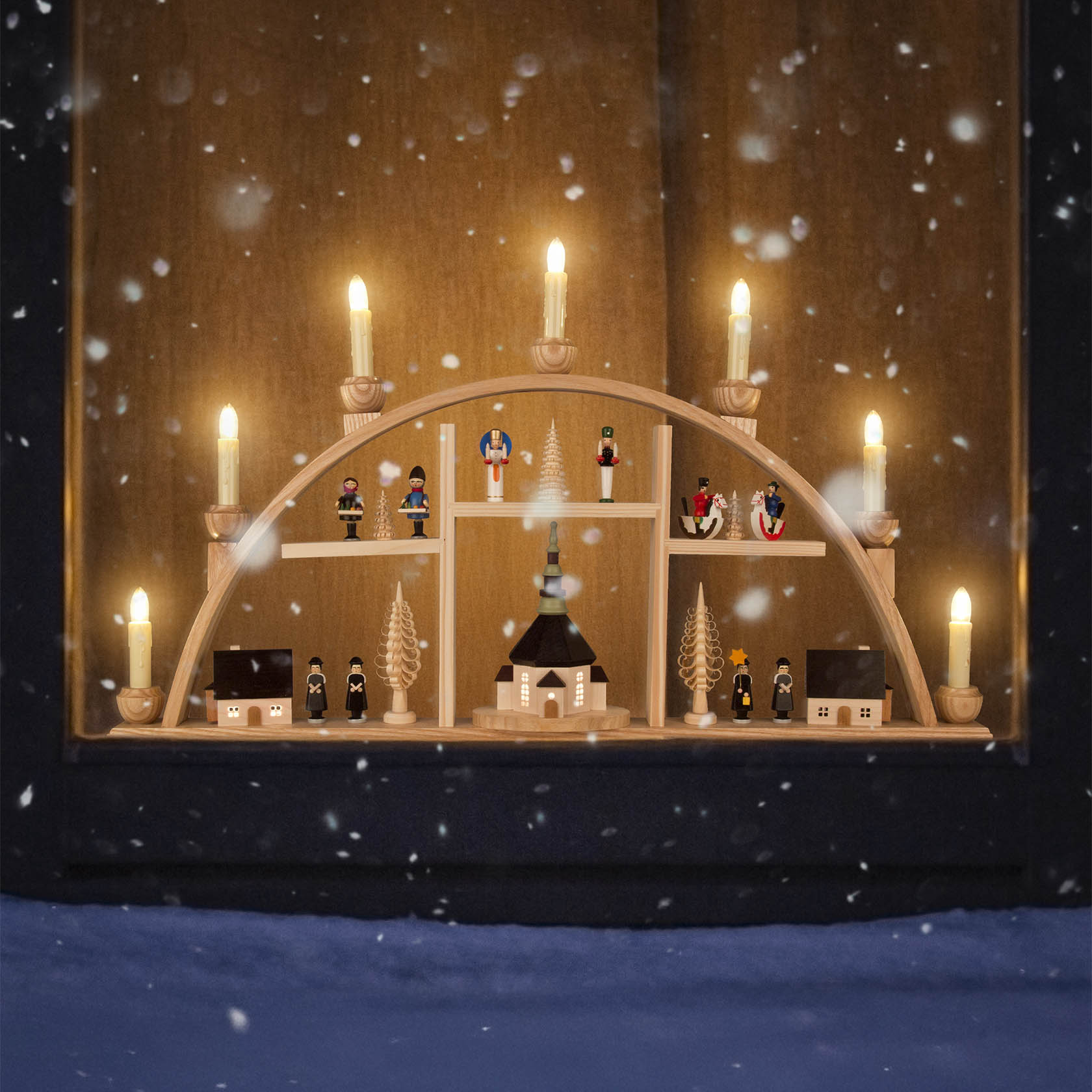 Ein Schwibbogen mit Seiffener Kirche, Kurrende-Sängern und Erzgebirgsfiguren, elektrisch beleuchtet, unter einem Fenster mit Schnee im Vordergrund.