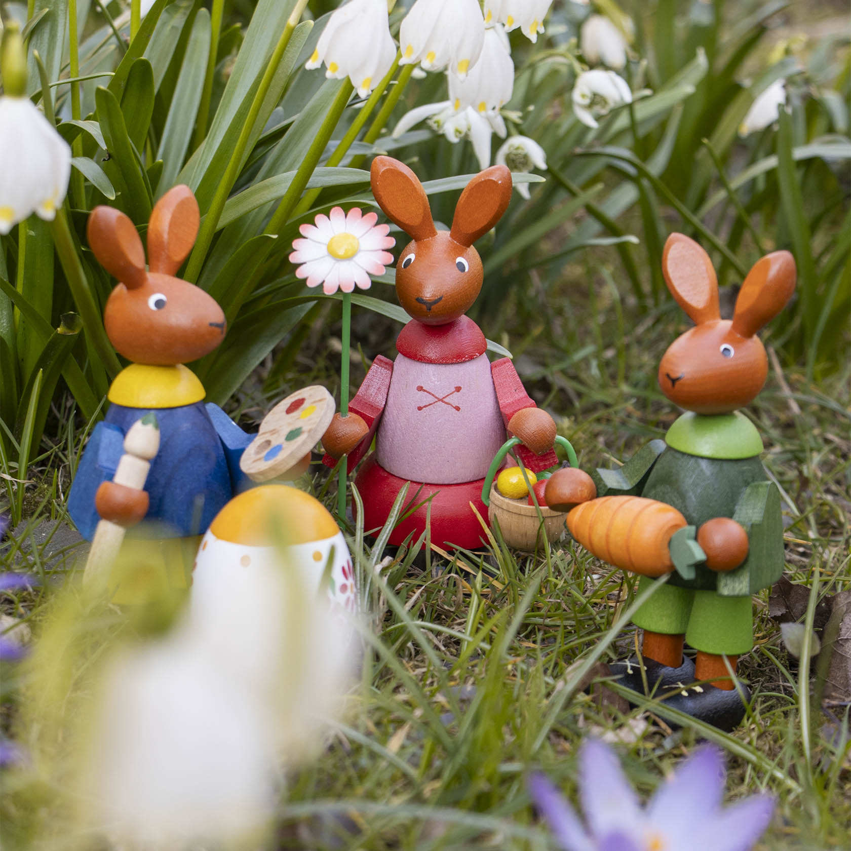 Vier hölzerne Osterhasenfiguren stehen in Gras zwischen Blumen. Einer hält eine Rübe, ein anderer einen Osterkorb.
