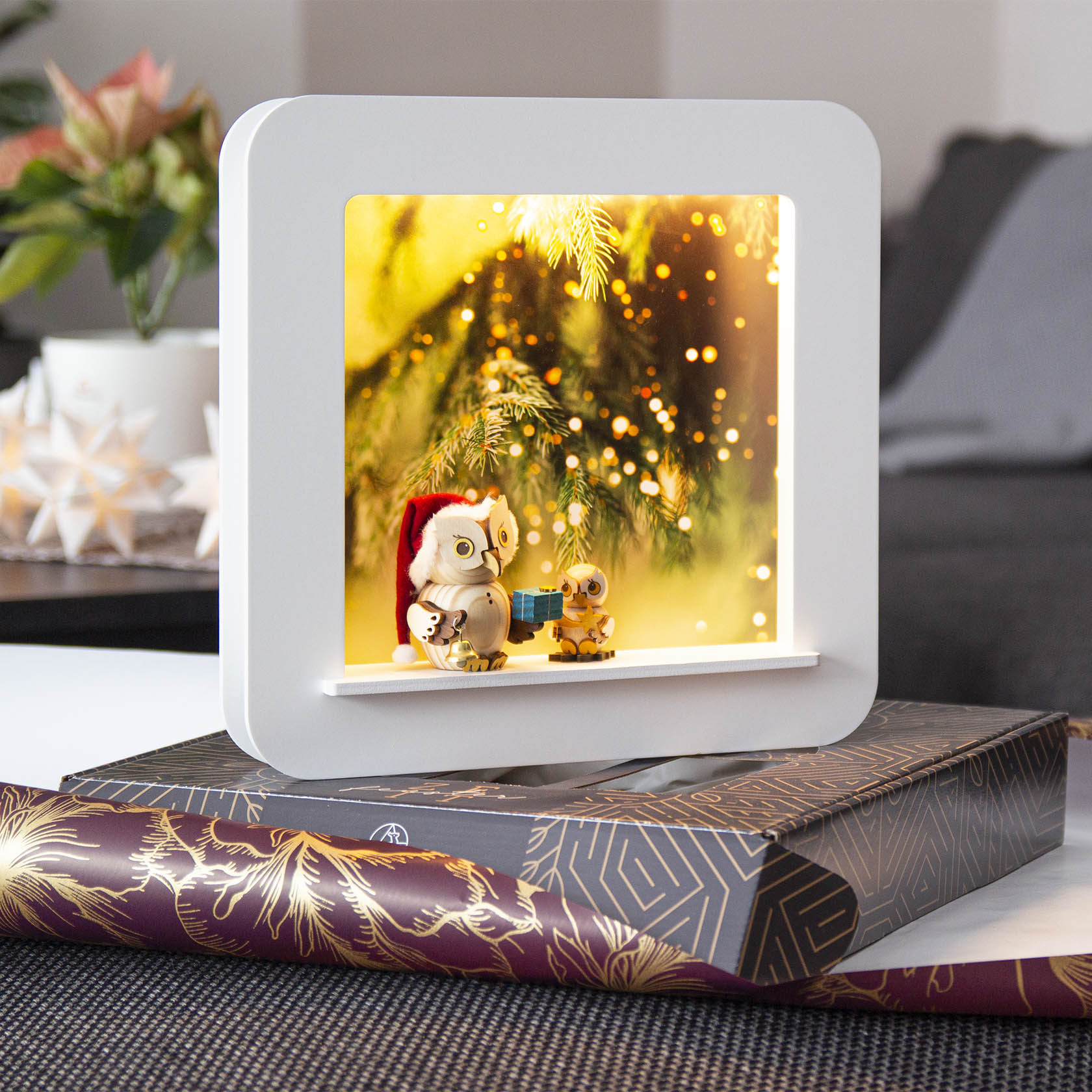 Zwei Mini-Eulenfiguren, eine mit Weihnachtsmannmütze und ein Geschenk in den Händen, vor einem weihnachtlich beleuchteten Hintergrund in einem weißen Rahmen.
