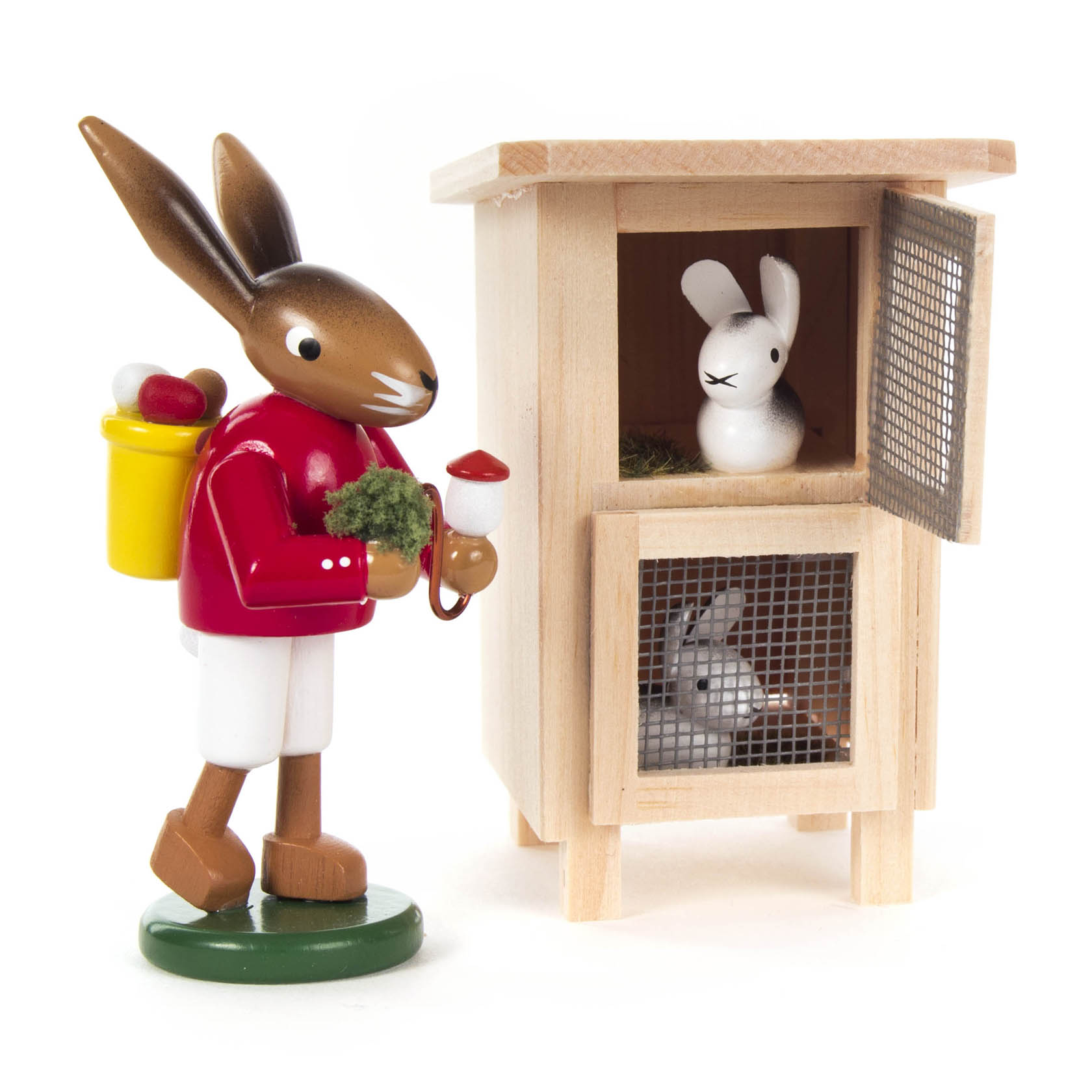 Holzfigur eines Hasen in rotem Mantel, der ein Osterei und Moos trägt, neben einem Holzhäschenstall mit zwei weiteren Hasenfiguren. im Dregeno Online Shop günstig kaufen