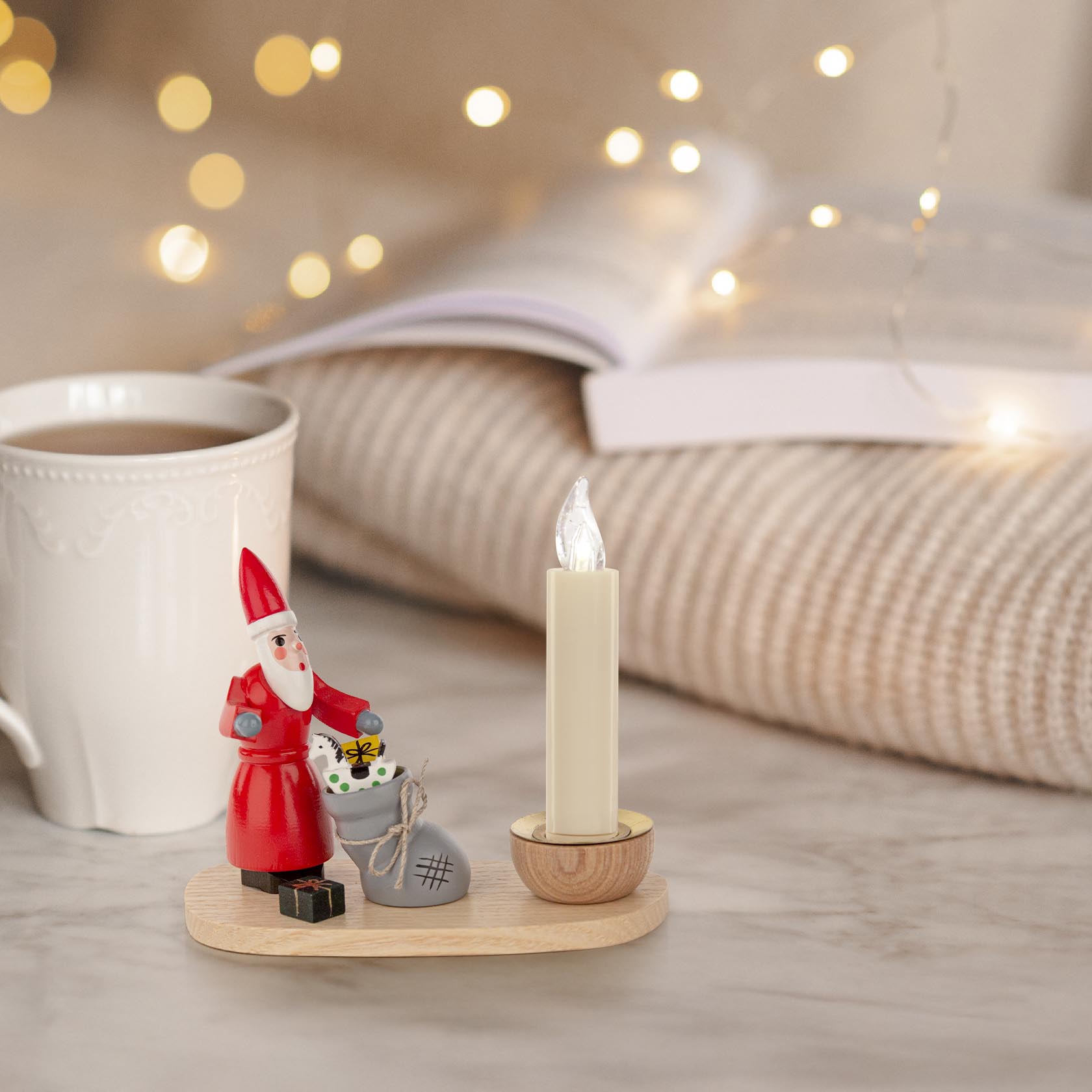 A decorative candle holder with a Santa Claus figurine holding a small Christmas tree, positioned next to a lit candle and a small gift box.