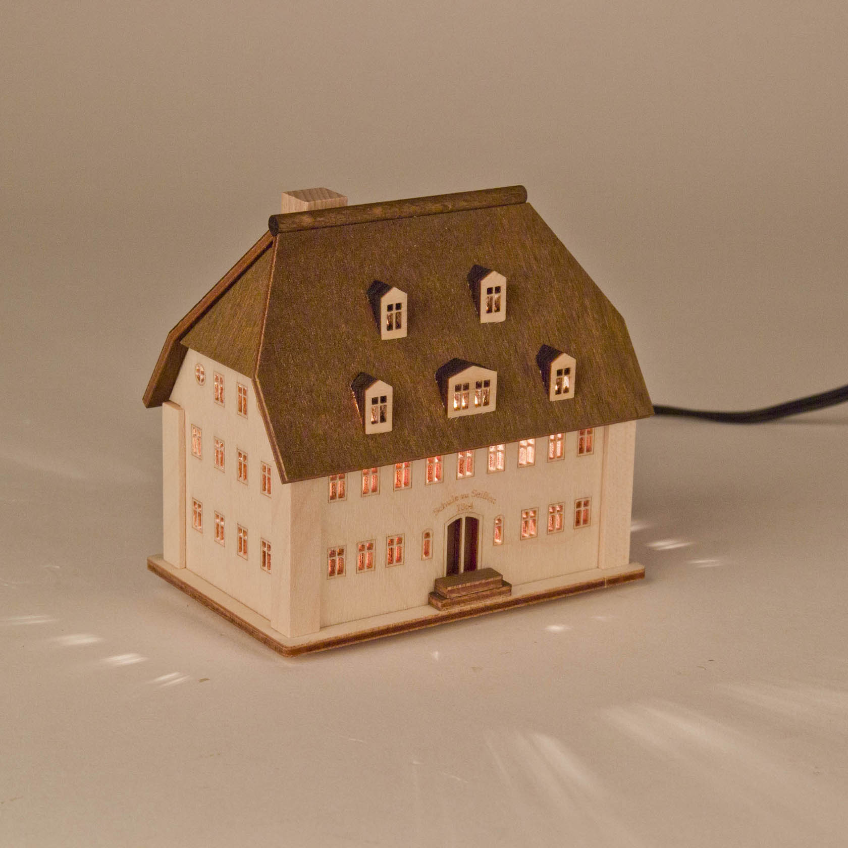 Ein beleuchtetes miniatur Lichterhaus in Form eines Schulgebäudes mit fünf Fenstern auf jedem der beiden Stockwerke.