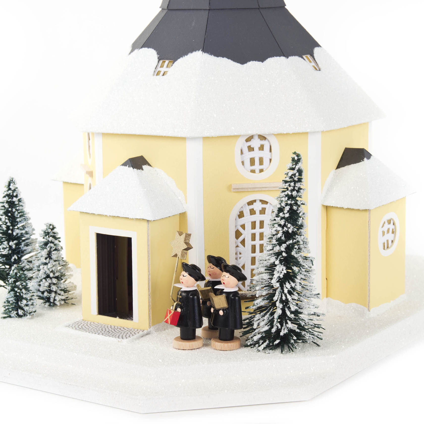 Detailreiche Seiffener Kirche mit schneebedecktem Dach, umgeben von Tannenbäumen. Zwei Kurrendesänger in traditioneller Kleidung stehen singend vor der Kirche.