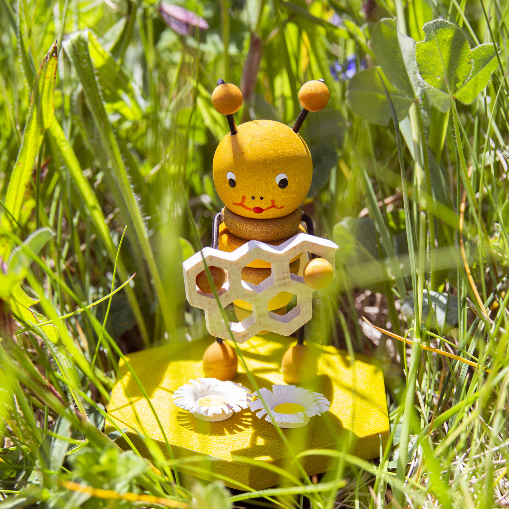 Holzfigur einer Biene hält eine stilisierte Honigwabe in den Händen, umgeben von Gras und Gänseblümchen.