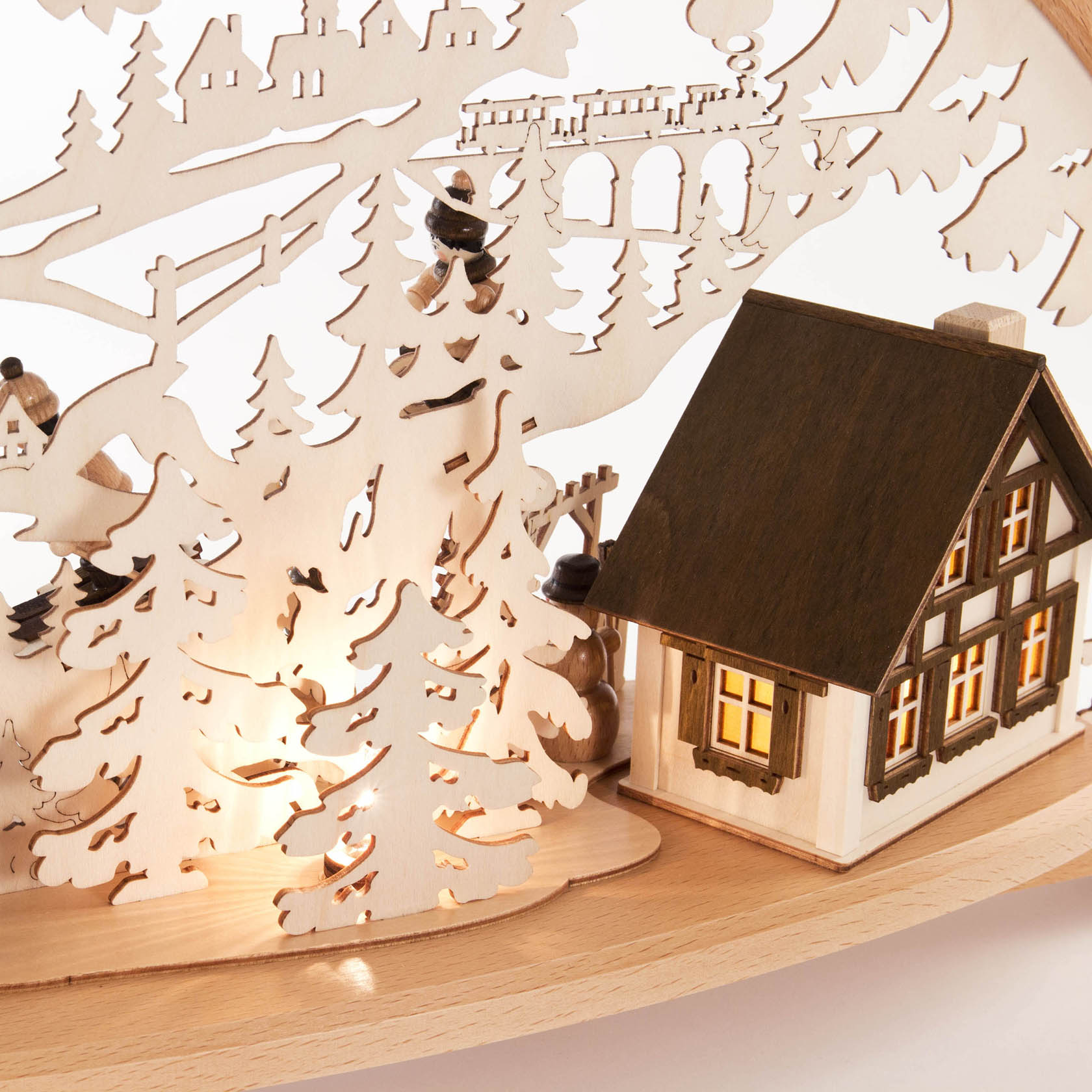 Ein Holz-Schwibbogen zeigt Wintersportler neben einem elektrisch beleuchteten Haus, umgeben von Bäumen, mit beleuchteten Fenstern und winterlicher Szene.