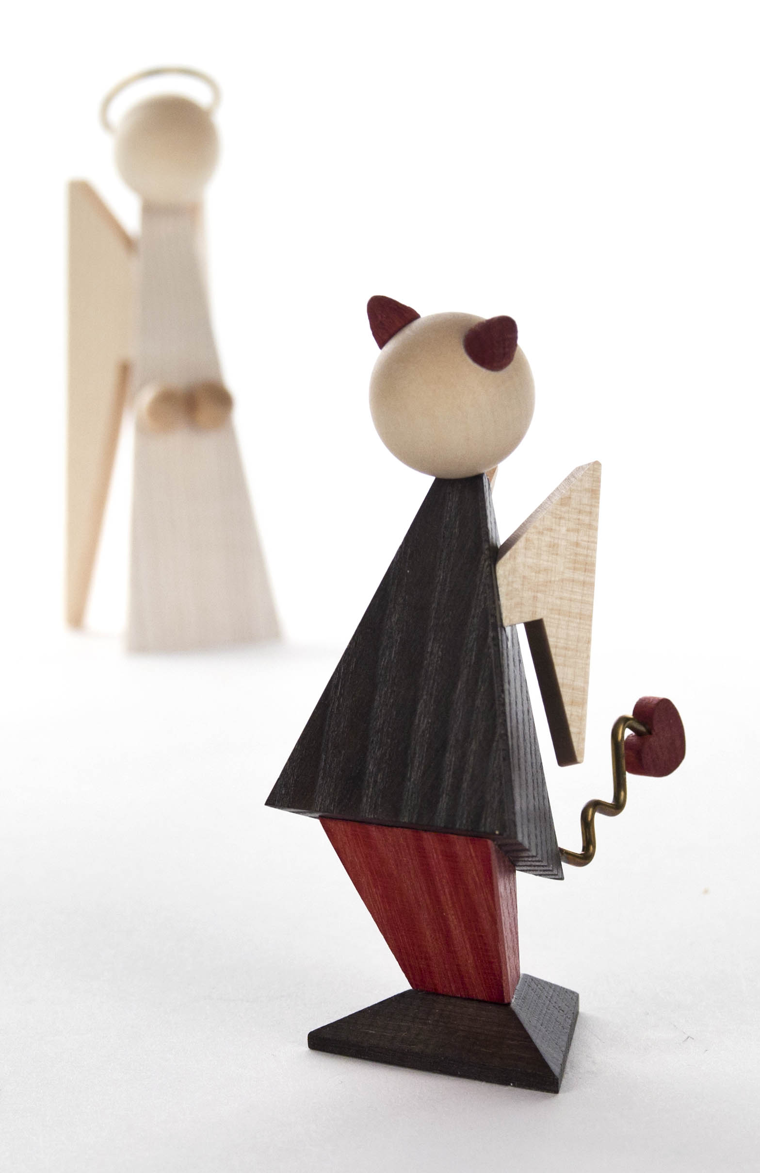 Holzfigur eines Teufels mit schwarzen Mantel, rotem Horn und spiralförmigem Schwanz, die von einer Engelsfigur im Hintergrund beobachtet wird.