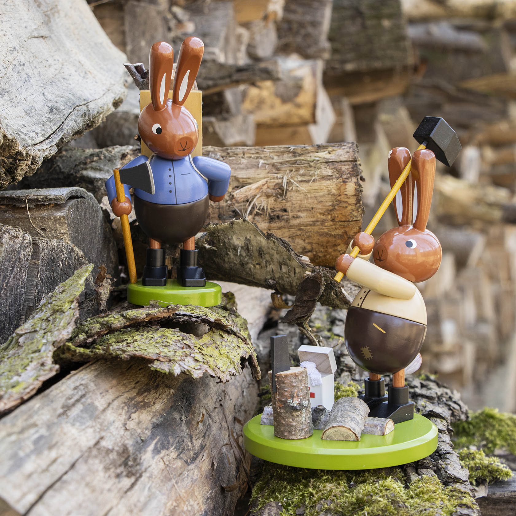Zwei Holzhasenfiguren. Der linke Hase trägt eine blaue Jacke und hält eine Axt, während der rechte Hase in heller Kleidung ein Holzscheit auf einem Hackklotz bearbeitet.
