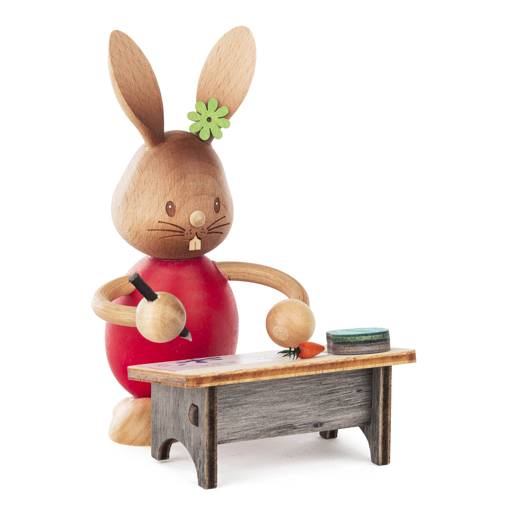 Holzfigur eines Hasen namens Stupsi in rotem Outfit mit Blume am Ohr, der an einem Tisch sitzt, eine Karotte betrachtet und einen Stift hält. im Dregeno Online Shop günstig kaufen