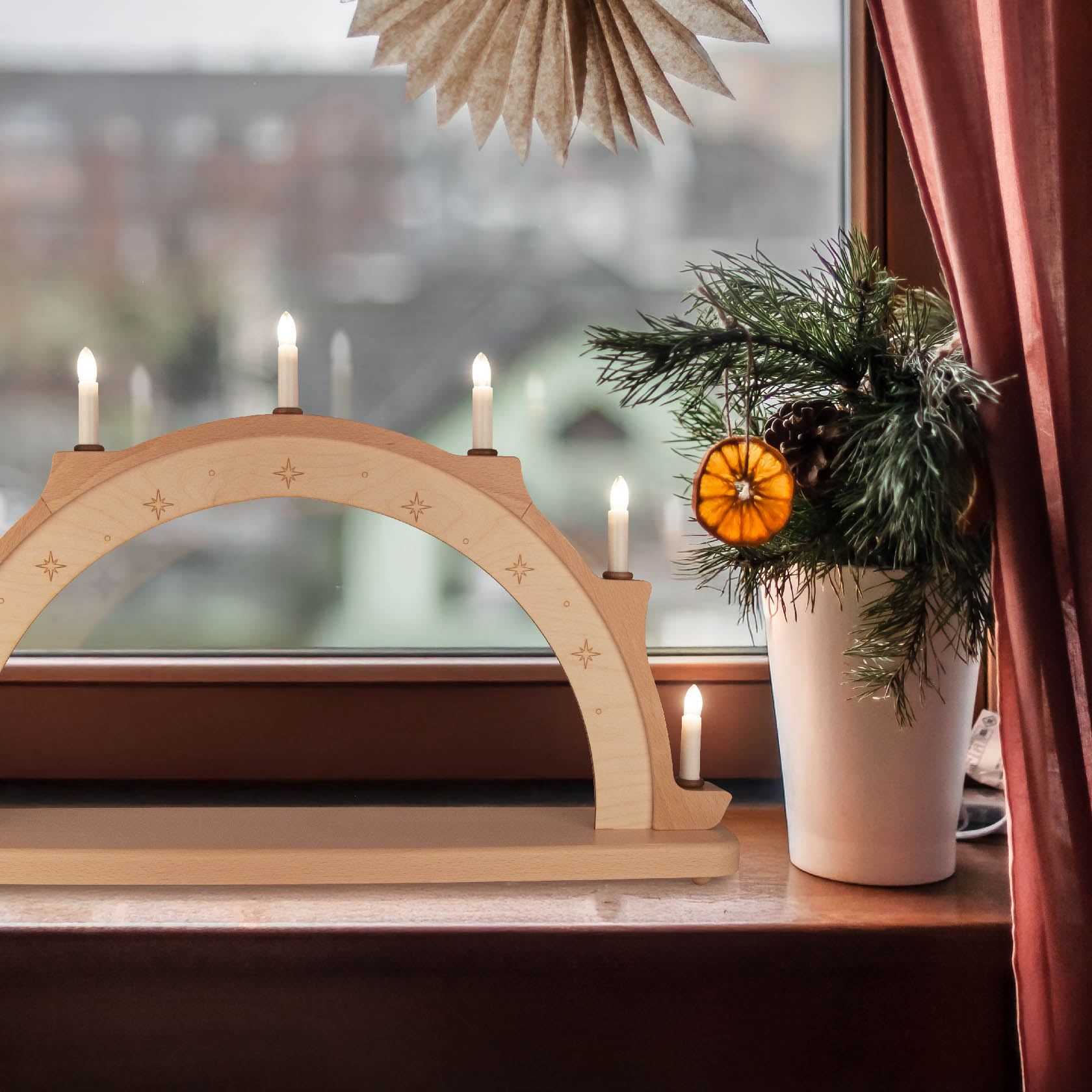 Schwibbogen aus hellem Holz mit Sternen graviert, sechs elektrisch leuchtende Kerzen, steht auf Fensterbank neben Topf mit Tannenzweig und getrockneter Orangenscheibe.