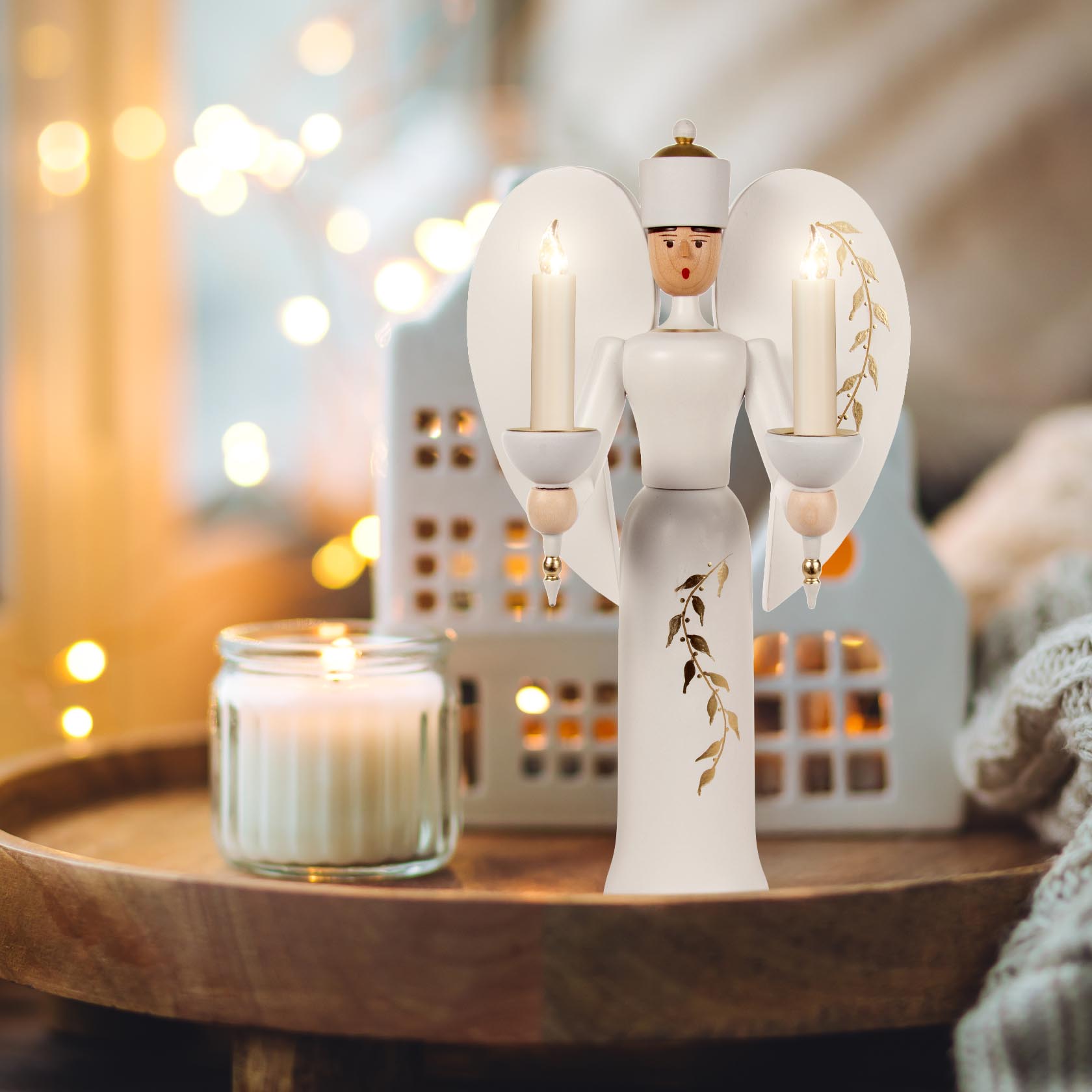 A white and gold angel figure designed to hold candles with floral patterns on its wings, standing on a wooden surface with a lit candle next to it.