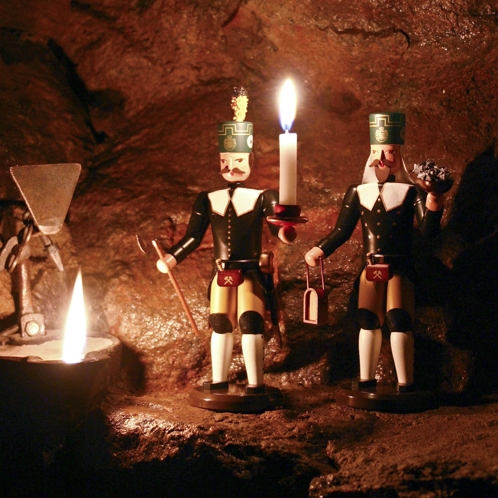 Zwei traditionelle Bergmannsfiguren aus Holz in Bergmannskleidung, eine hält einen Kerzenhalter mit brennender Kerze, die andere Blumen.