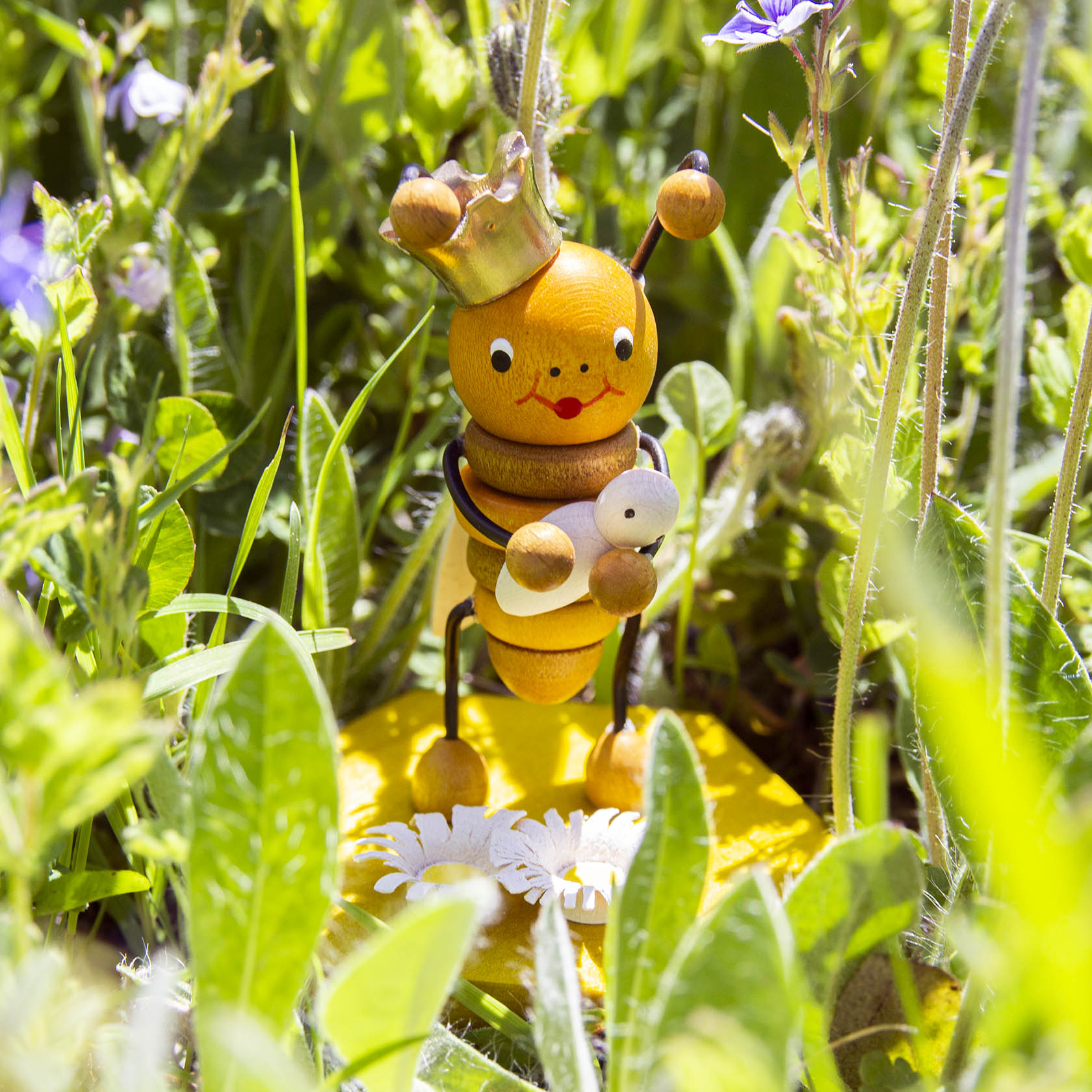 Holzfigur einer Bienenkönigin mit Krone, die ein Baby in den Armen hält, umgeben von grünen Pflanzen.
