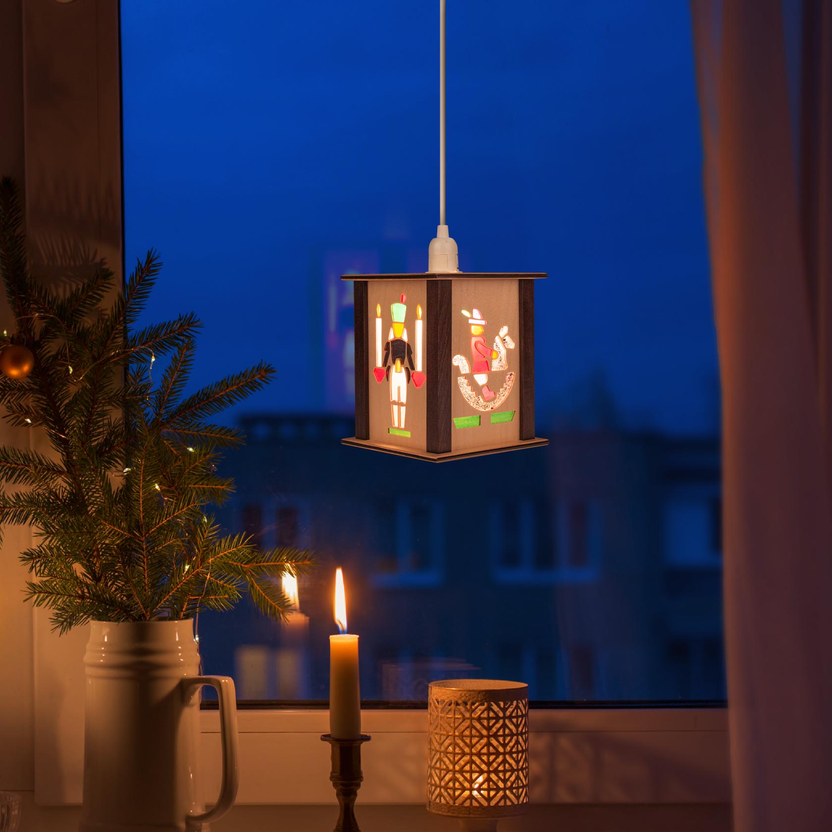 Papierlaterne mit aufgemaltem Reiter und Pferd hängt an einer Leuchte. Daneben ein Kerzenlicht und ein kleiner Tannenzweig auf der Fensterbank.