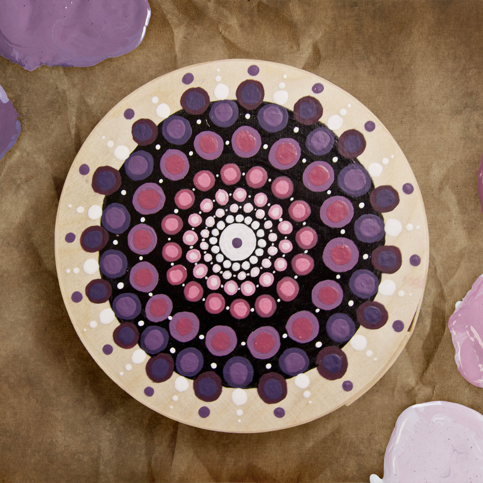Runde Spanschachtel mit einem Mandala-Design aus Punkten in verschiedenen Lilatönen und Weiß, auf einem braunen Untergrund.