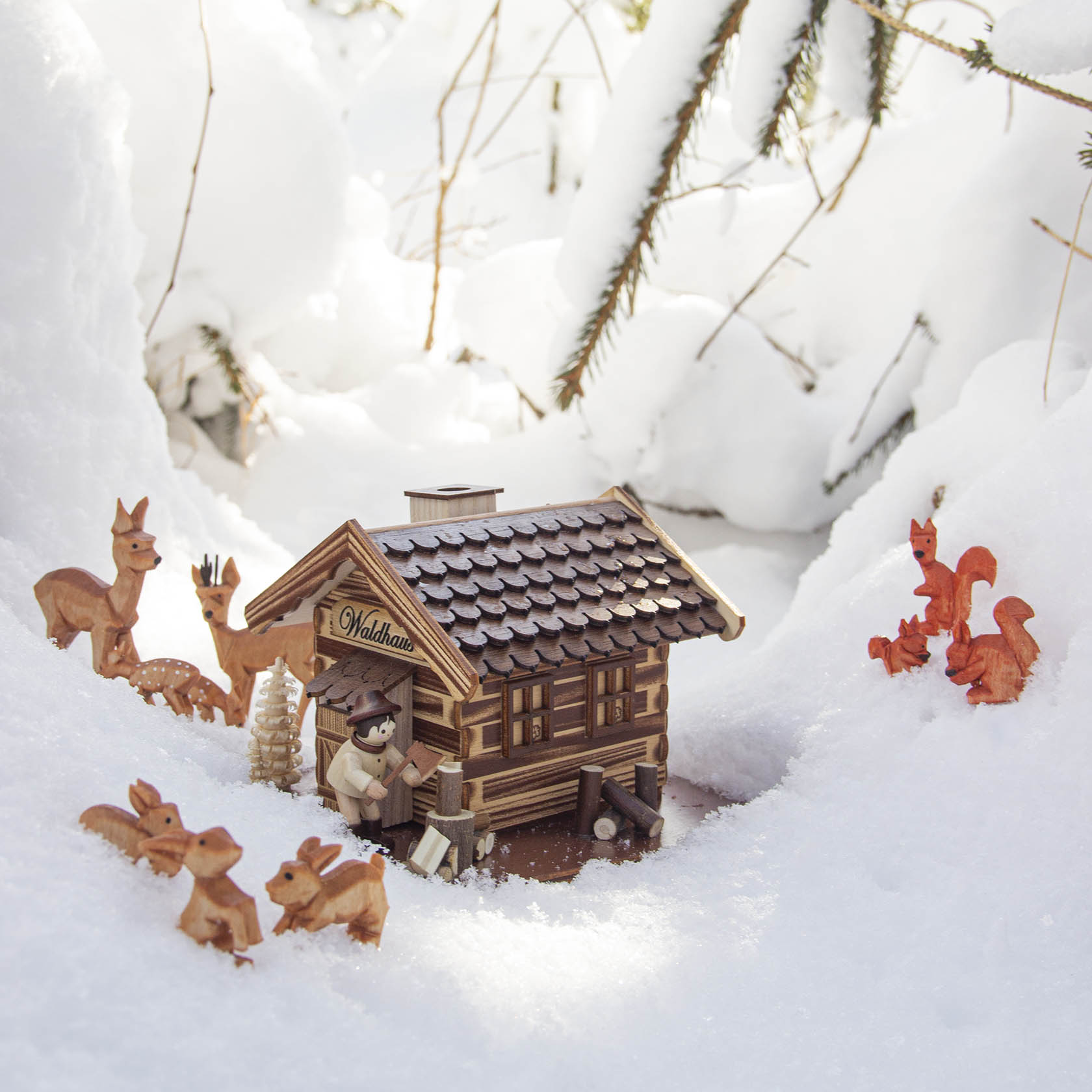 Holzhütte mit Schild 'Waldhaus', geschnitzte Figuren von zwei Eichhörnchen, einem Mann in traditioneller Kleidung und vier Rehen, umgeben von Schnee.