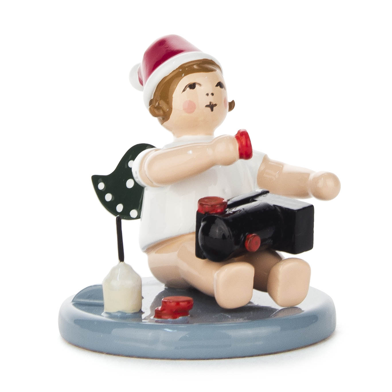 Ein sitzender, hölzerner Engel mit grünen Flügeln, einem roten Hut, hält eine schwarze Eisenbahn und ist neben einem Leimtopf und roter Farbe positioniert. im Dregeno Online Shop günstig kaufen