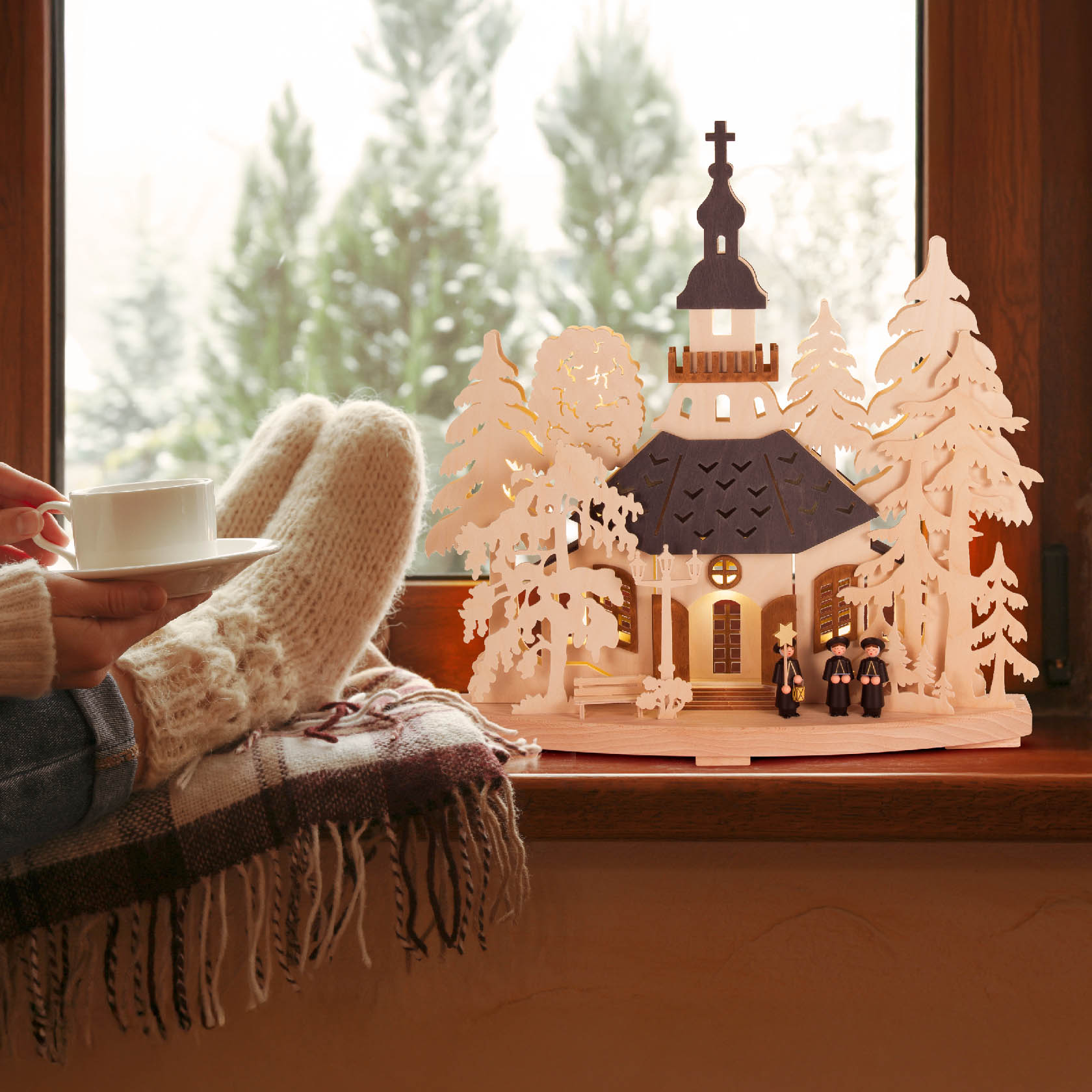 Ein Schwibbogen mit dem Motiv 'Seiffener Kirchgang', zeigt eine Holzkirche und Figuren, von innen elektrisch beleuchtet. Im Hintergrund sitzt eine Person mit Wollsocken.
