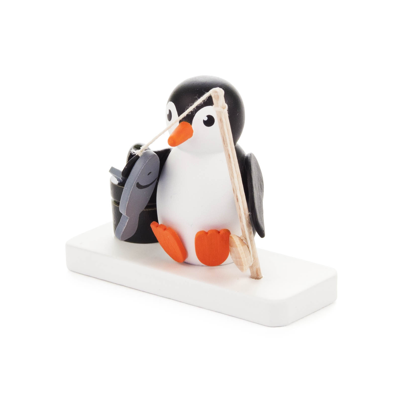 Eine kleine Holzfigur zeigt einen niedlichen Pinguin, der mit einer Angelrute einen Fisch aus einem Eimer fängt. Die Figur ist auf einem weißen Sockel montiert.