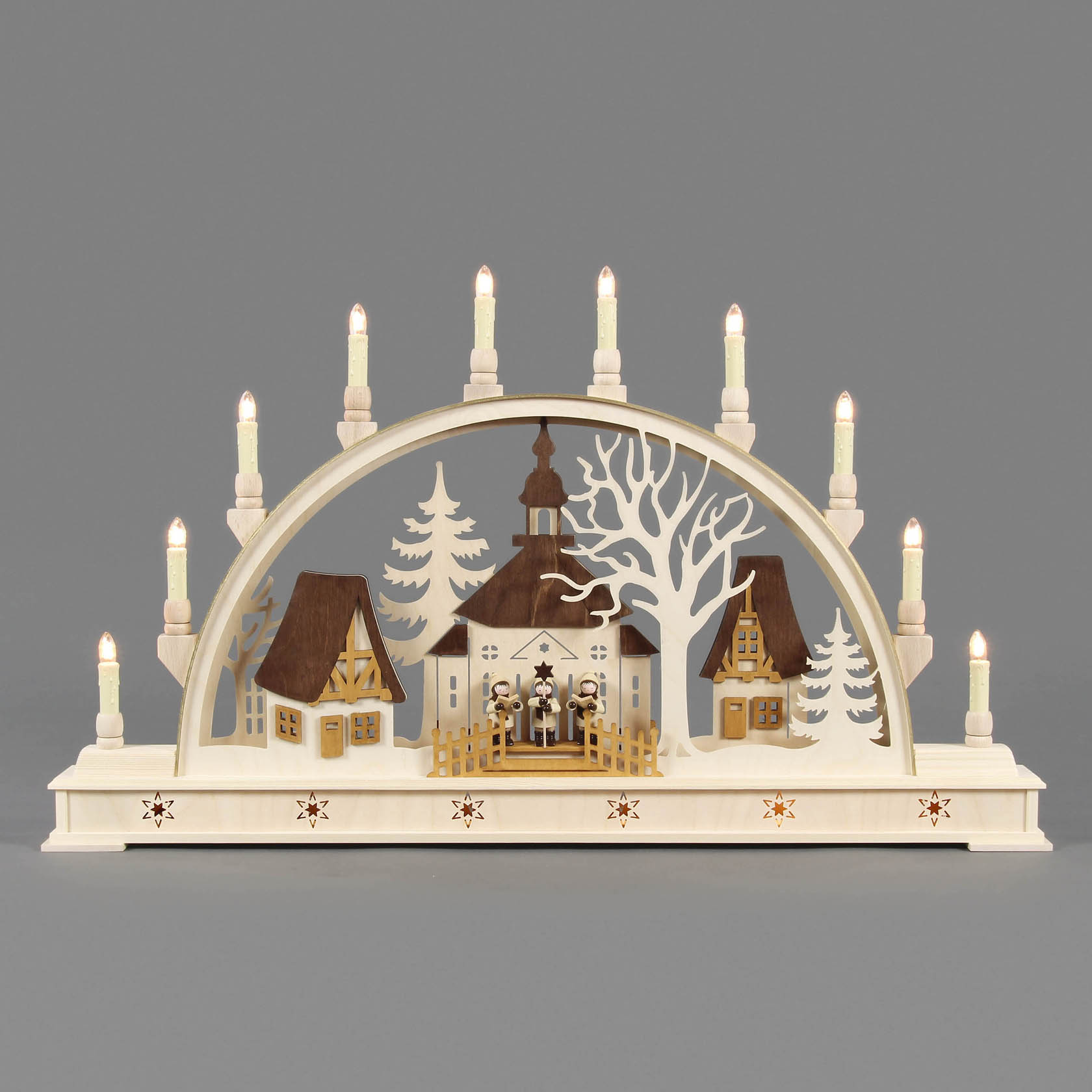 Ein Schwibbogen zeigt ein traditionelles, elektrisch beleuchtetes Seiffener Dorf mit Kurrendefiguren zwischen zwei verzierten Häusern und einem zentralen Baum.