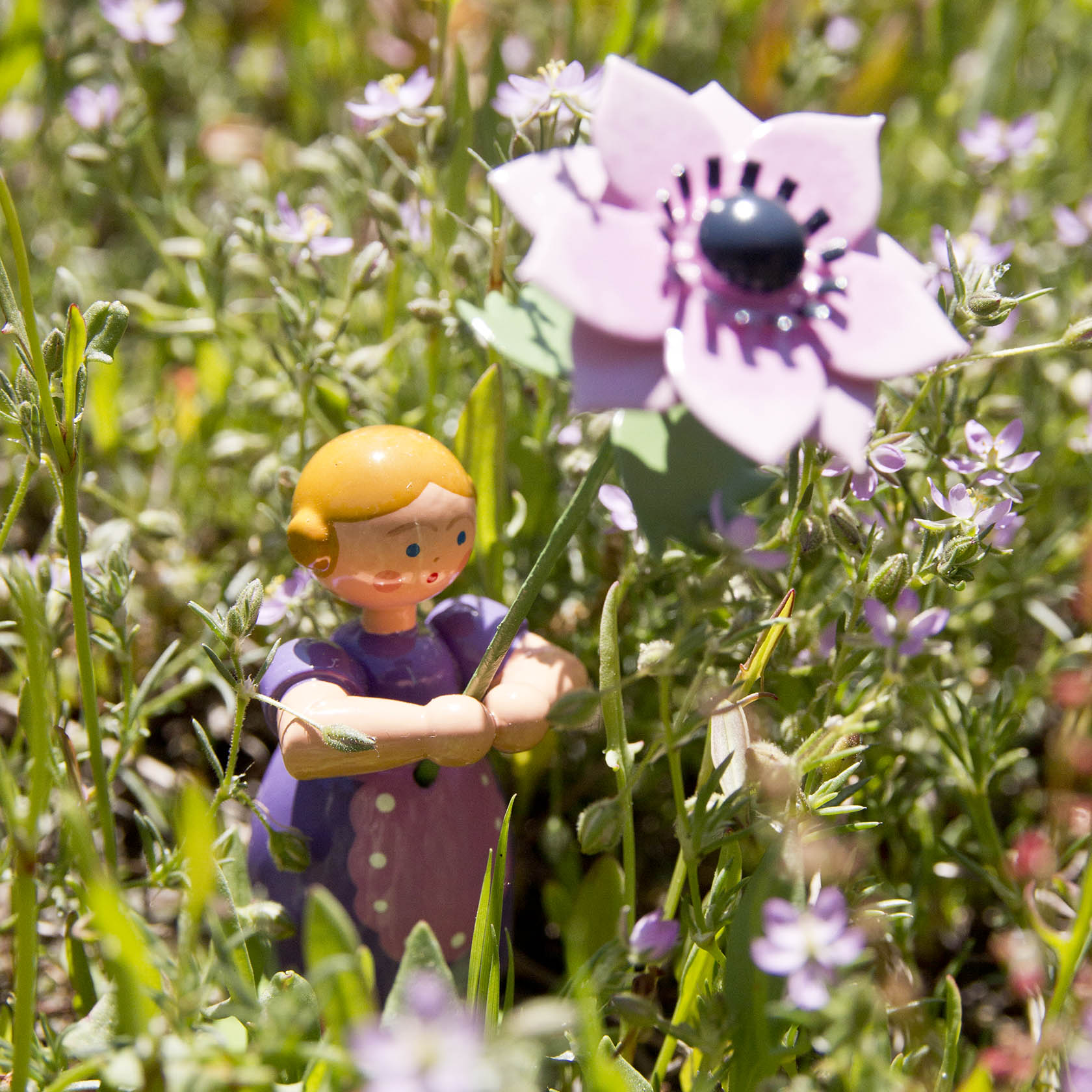 Eine kleine Holzfigur eines Mädchens mit orangeblonden Haaren hält eine große Anemone. Die Figur steht inmitten von Gras und kleinen violetten Blumen.
