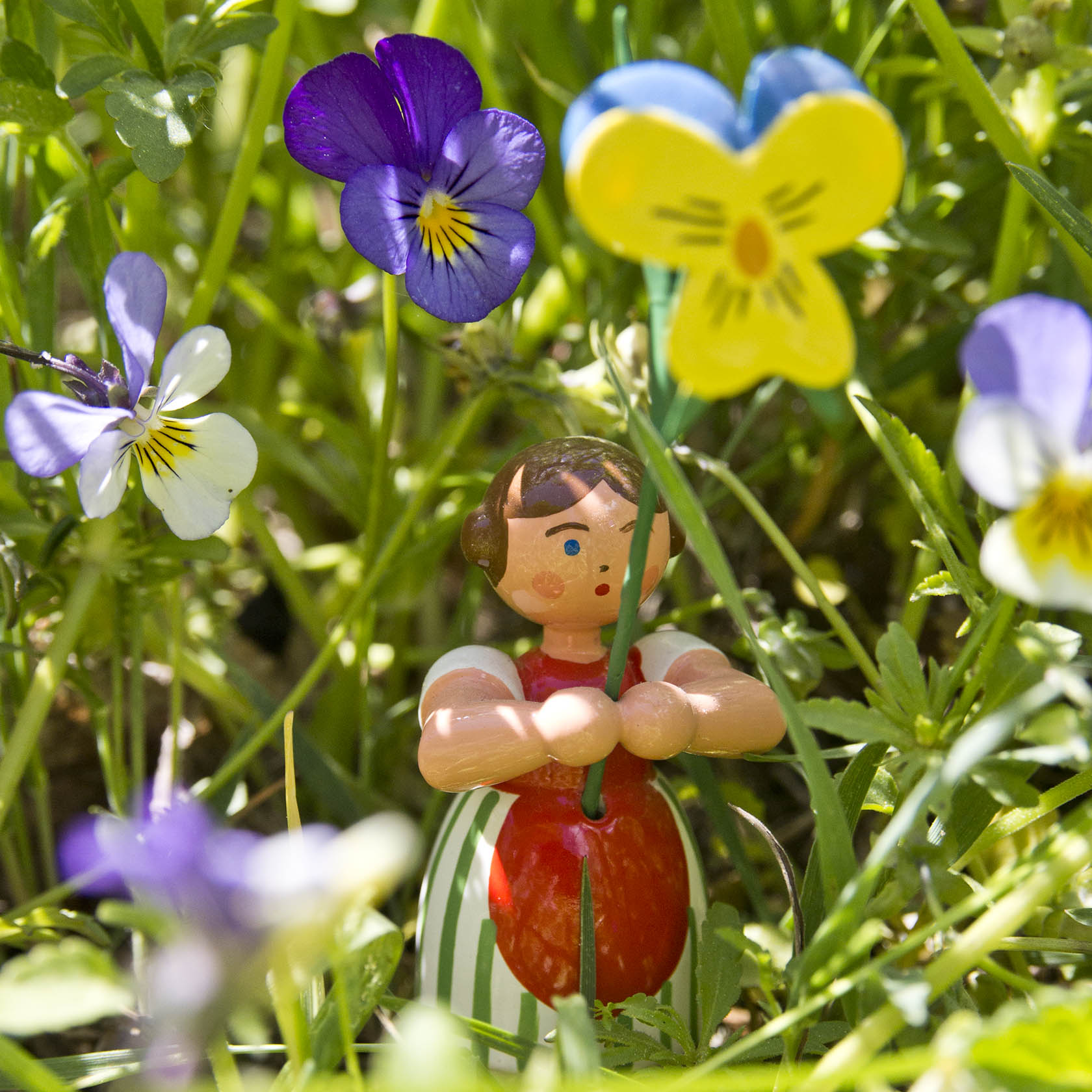 Eine hölzerne Blumenmädchenfigur in einem gestreiften Kleid hält ein gelbes Blütenblatt, umgeben von blühenden violetten Stiefmütterchen im Gras.