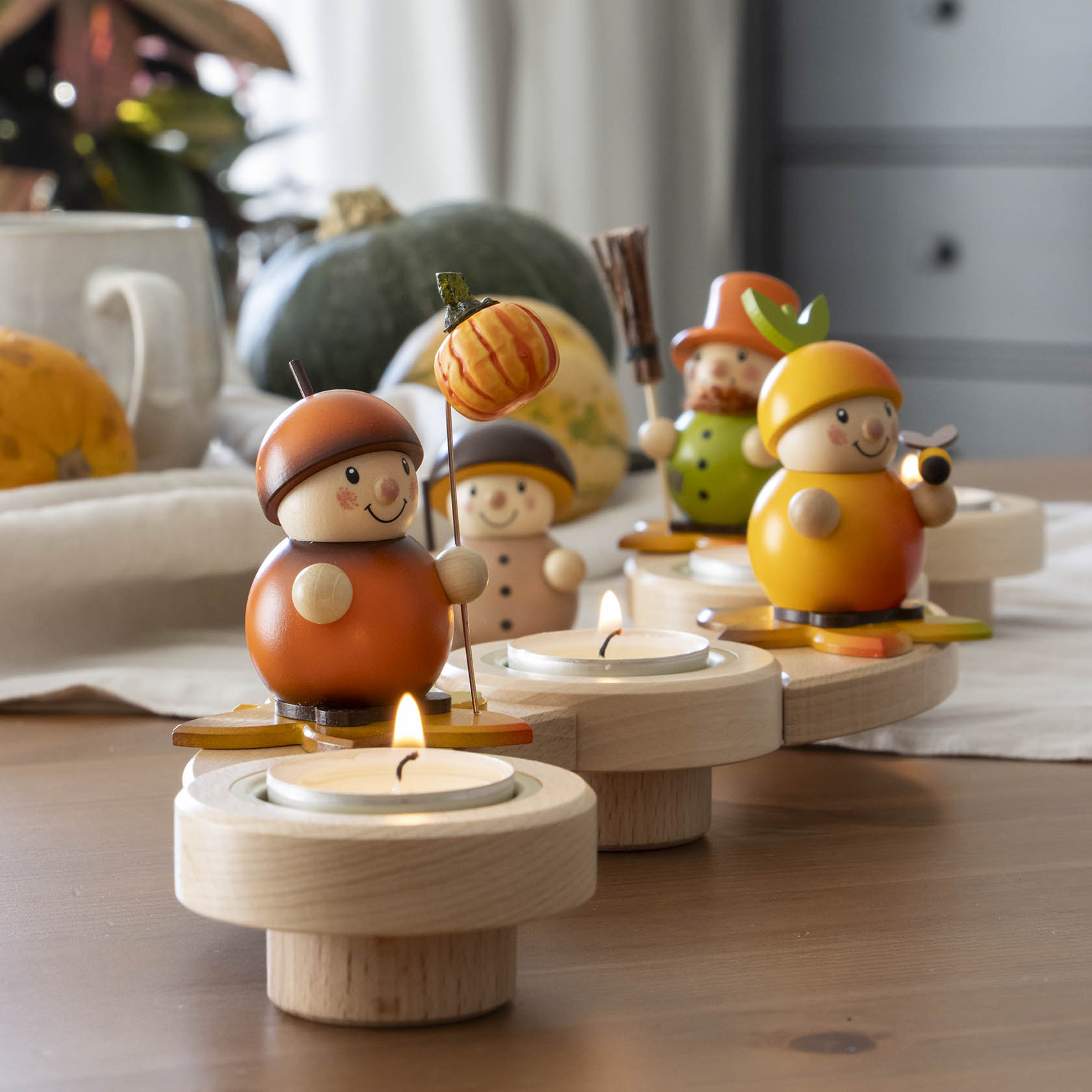 Zwei hölzerne Vogelscheuchenfiguren in Herbstfarben auf einem Vario-Leuchter. Eine hält einen Besen, die andere einen Kürbis-Stecker.