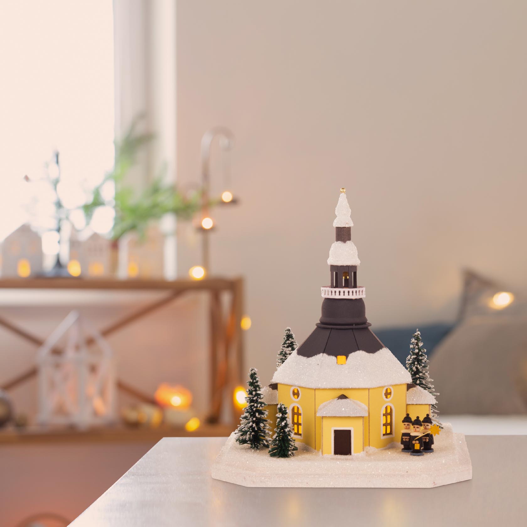 Das Lichterhaus Seiffener Kirche: Ein kleines Modell einer Kirche in Gelb mit schwarzem Dach, bedeckt von Schneeimitation, umgeben von Miniaturtannen und Figuren.