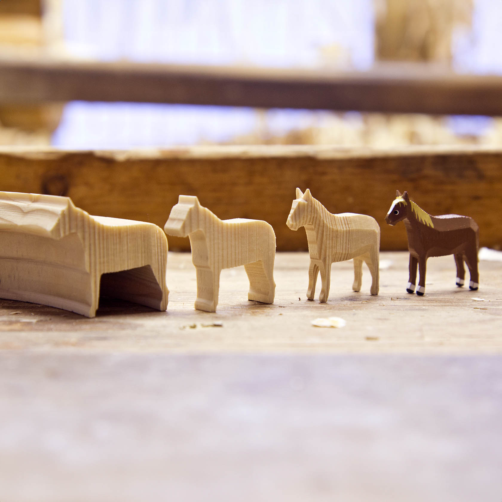 Vier kleine Holztiere, darunter zwei Pferde und ein Esel, stehen aufgereiht vor einem umzäunten Bereich auf einem Holztisch.