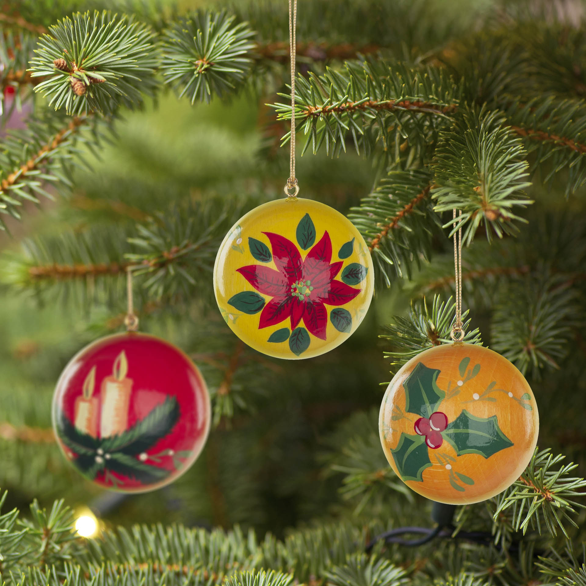 Drei Weihnachtsbaumkugeln mit Motiven: rote Kerzen auf einer Tanne, rote Blume auf gelbem Hintergrund, Stechpalmenzweig auf orangem Hintergrund.