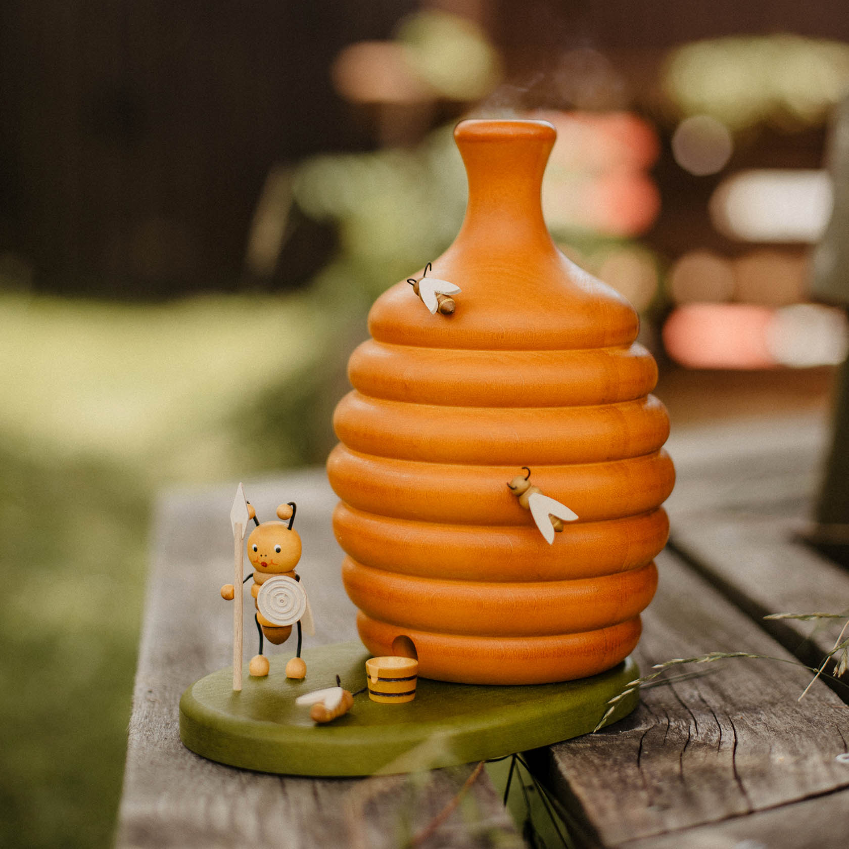 Dekorativer Holz-Bienenstock mit Bienenfiguren und Räucherfunktion, auf einem Holzsockel stehend. Kleine Bienenfiguren und Zubehör wie ein Honigeimer sind dabei.