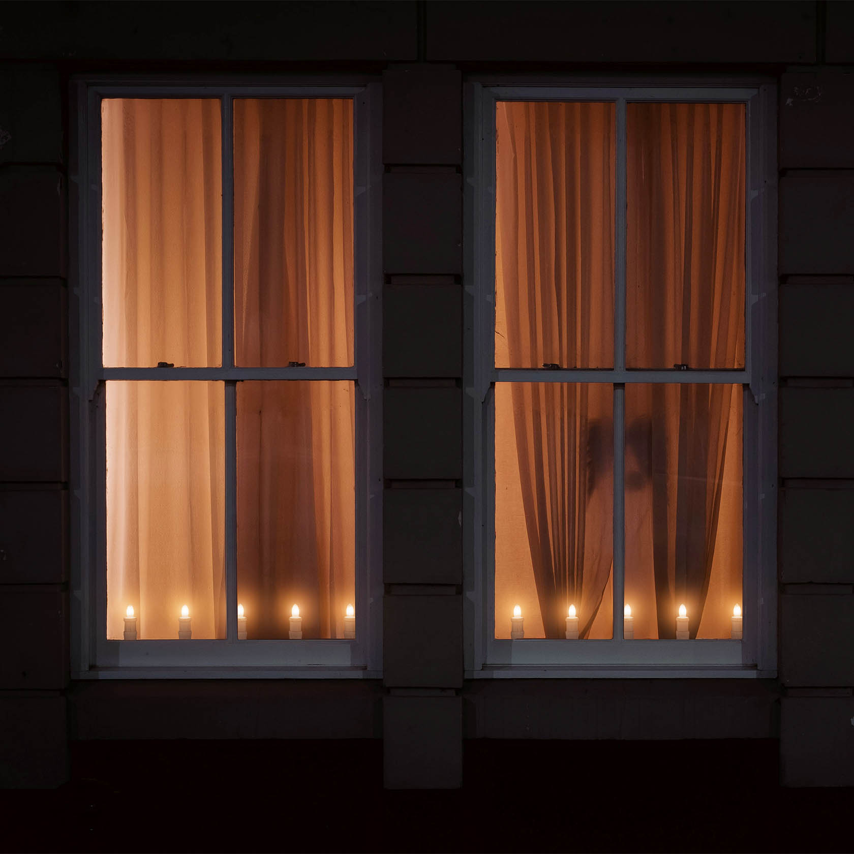 Zwei Fenster mit langen Vorhängen, durch die eine Lichterleiste mit fünf leuchtenden Riffelkerzen sichtbar ist.