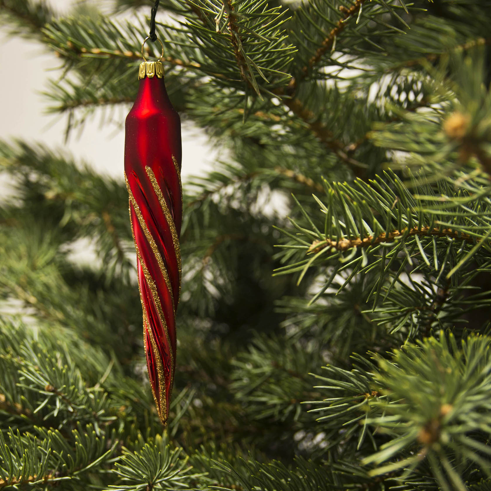 Roter, matt schimmernder Eiszapfen-Christbaumschmuck mit Goldglimmer, hängt an einem Tannenzweig.